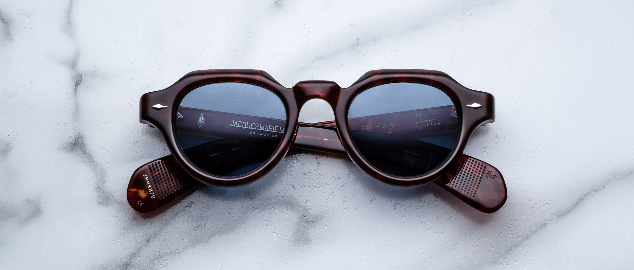 Jacques Marie Mage Kellerman round tortoiseshell sunglasses with blue-tinted lenses rest upside down on a white marble surface, temples folded neatly.