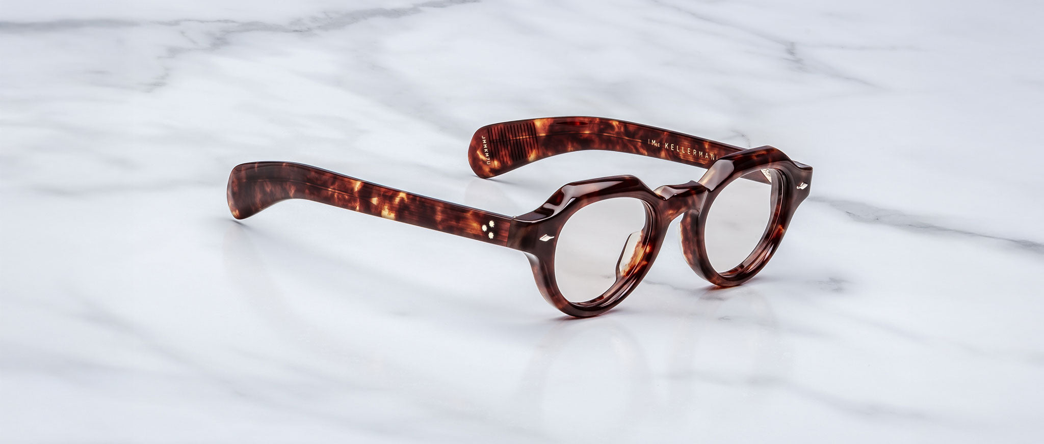 A pair of Jacques Marie Mage Kellerman round, thick-framed tortoiseshell eyeglasses rests on a white marble surface.