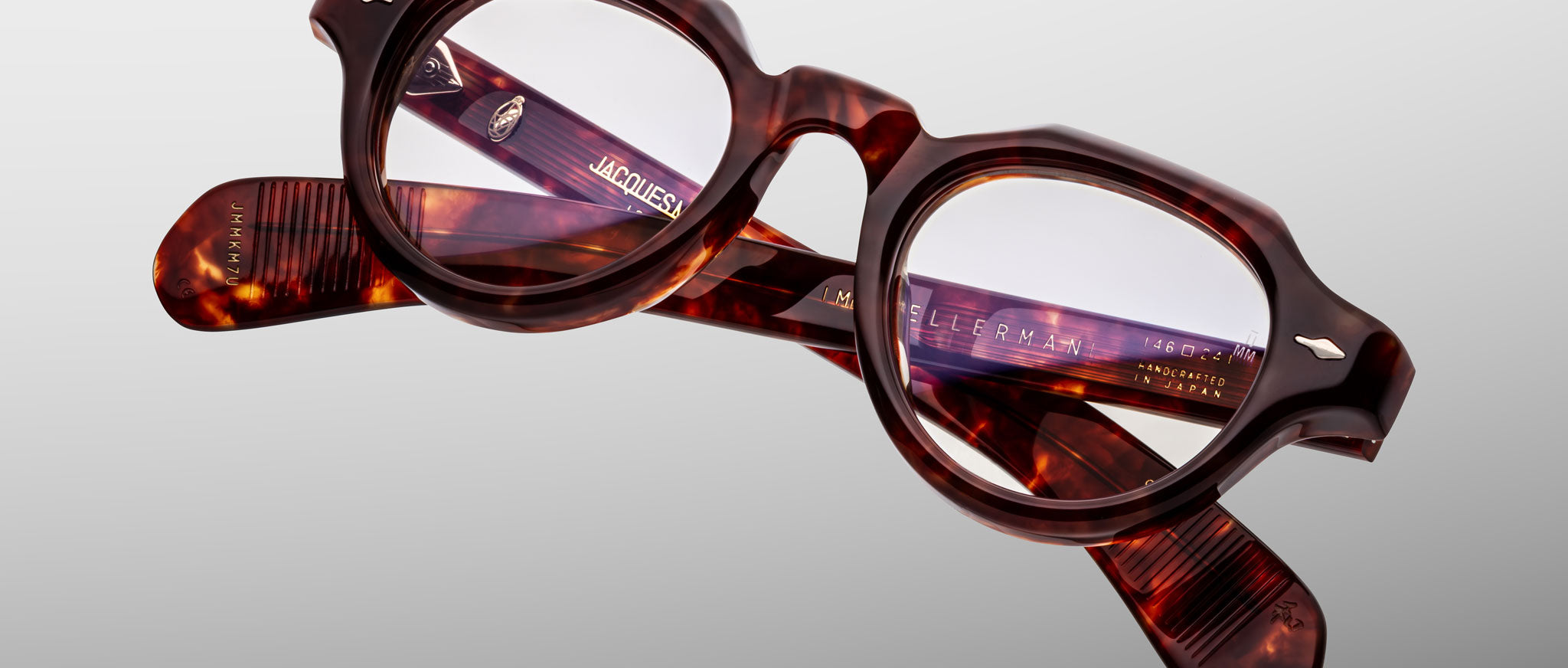 A pair of Jacques Marie Mage Kellerman eyeglasses featuring brown tortoiseshell, round lenses, and thick frames rests on a light gray background, with the brand name and details shown on the inside of the arms.