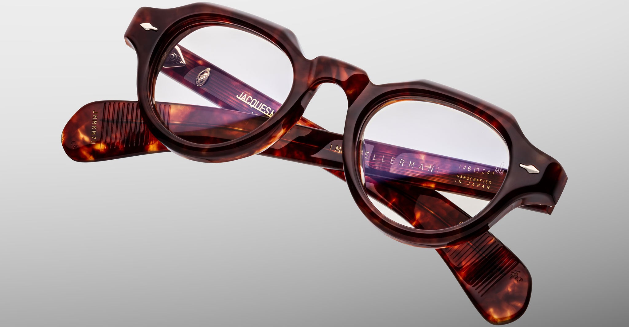 A pair of Jacques Marie Mage Kellerman eyeglasses featuring brown tortoiseshell, round lenses, and thick frames rests on a light gray background, with the brand name and details shown on the inside of the arms.