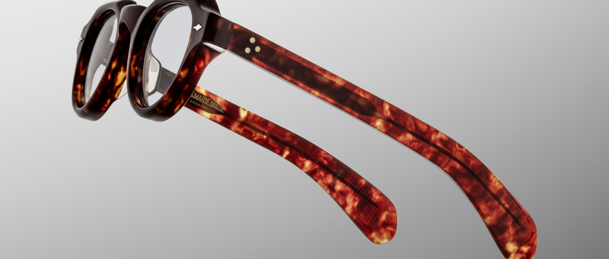The Jacques Marie Mage Kellerman features round lenses and thick tortoiseshell frames, shown at an angle to highlight the distinctive arms and side details against a plain grey background.