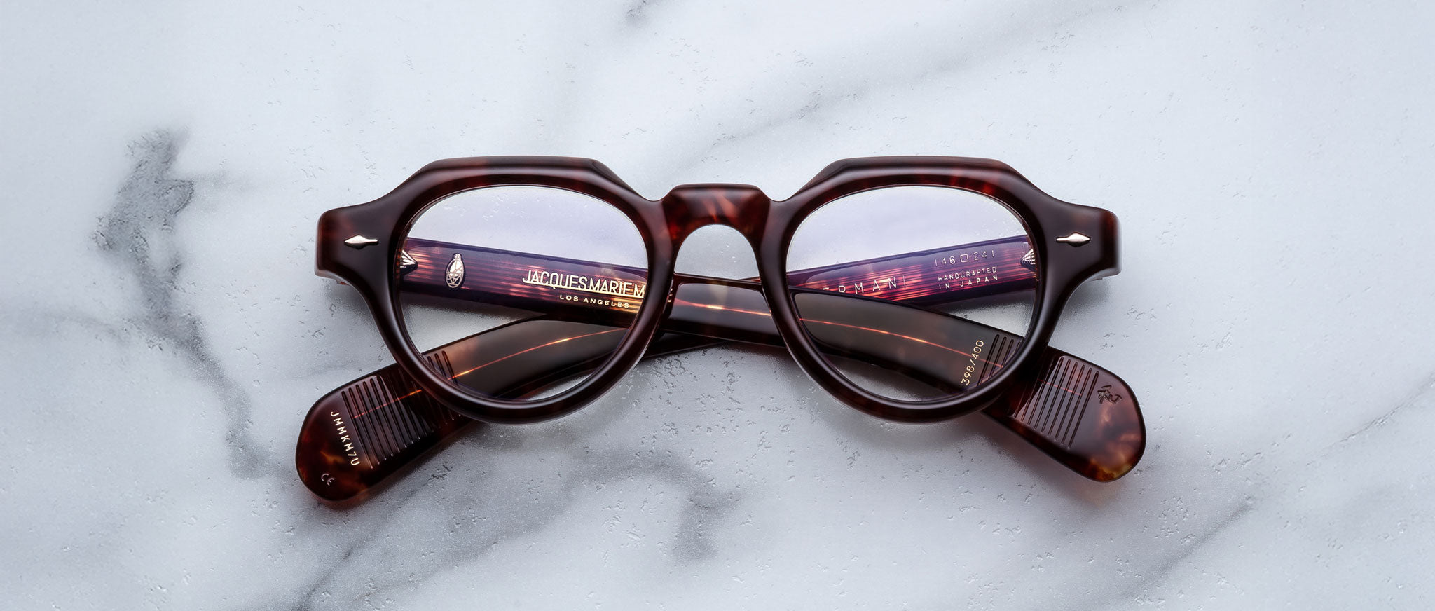A pair of Jacques Marie Mage Kellerman round, brown tortoiseshell eyeglasses rests on a white marble surface with the arms crossed beneath the lenses.