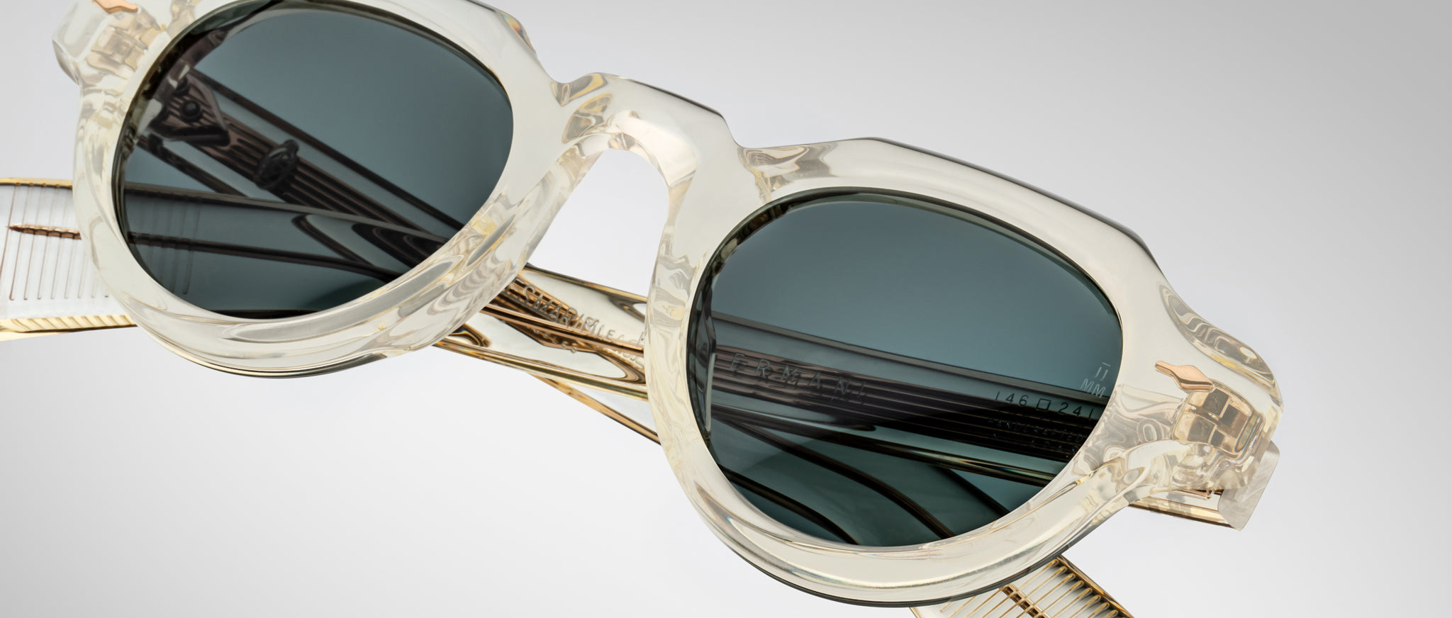 Jacques Marie Mage Kellerman clear-framed sunglasses with dark tinted lenses rest on a reflective surface, casting distinct reflections of the frames and lenses against a soft light gradient background.