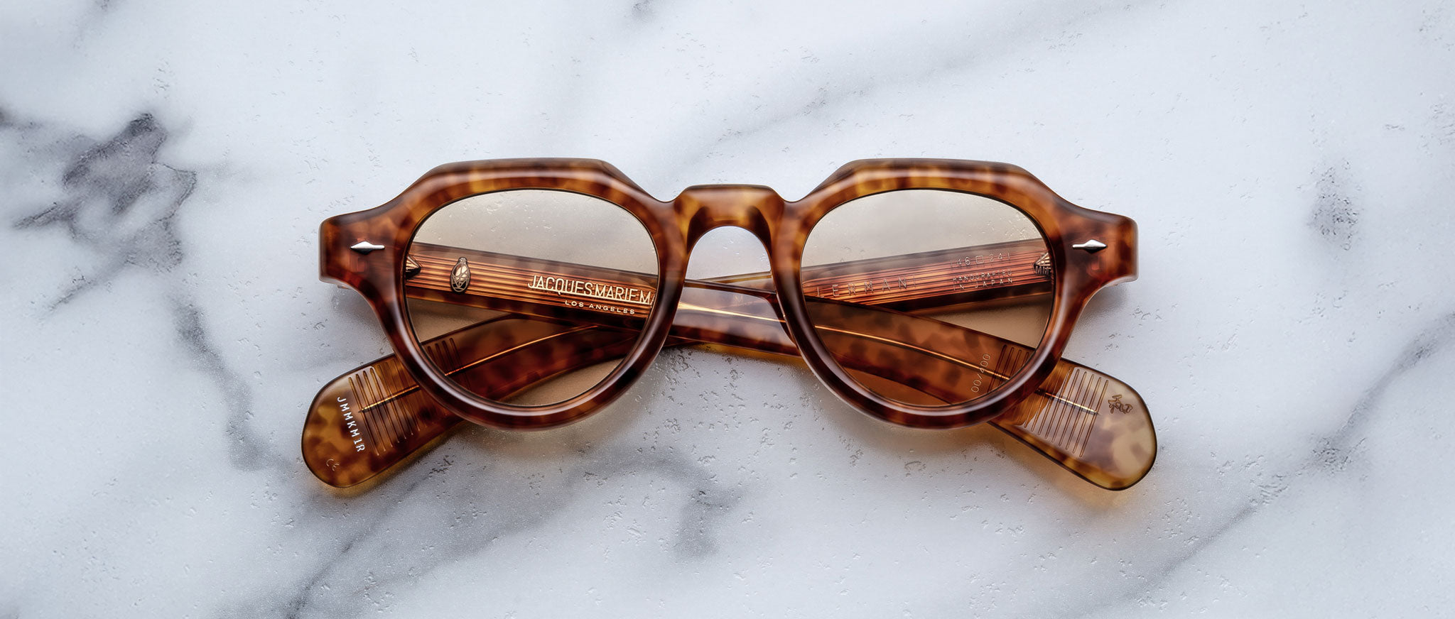 Jacques Marie Mage Kellerman brown tortoiseshell sunglasses with round lenses are folded and resting on a white marble surface, with the arms visible through the lenses.