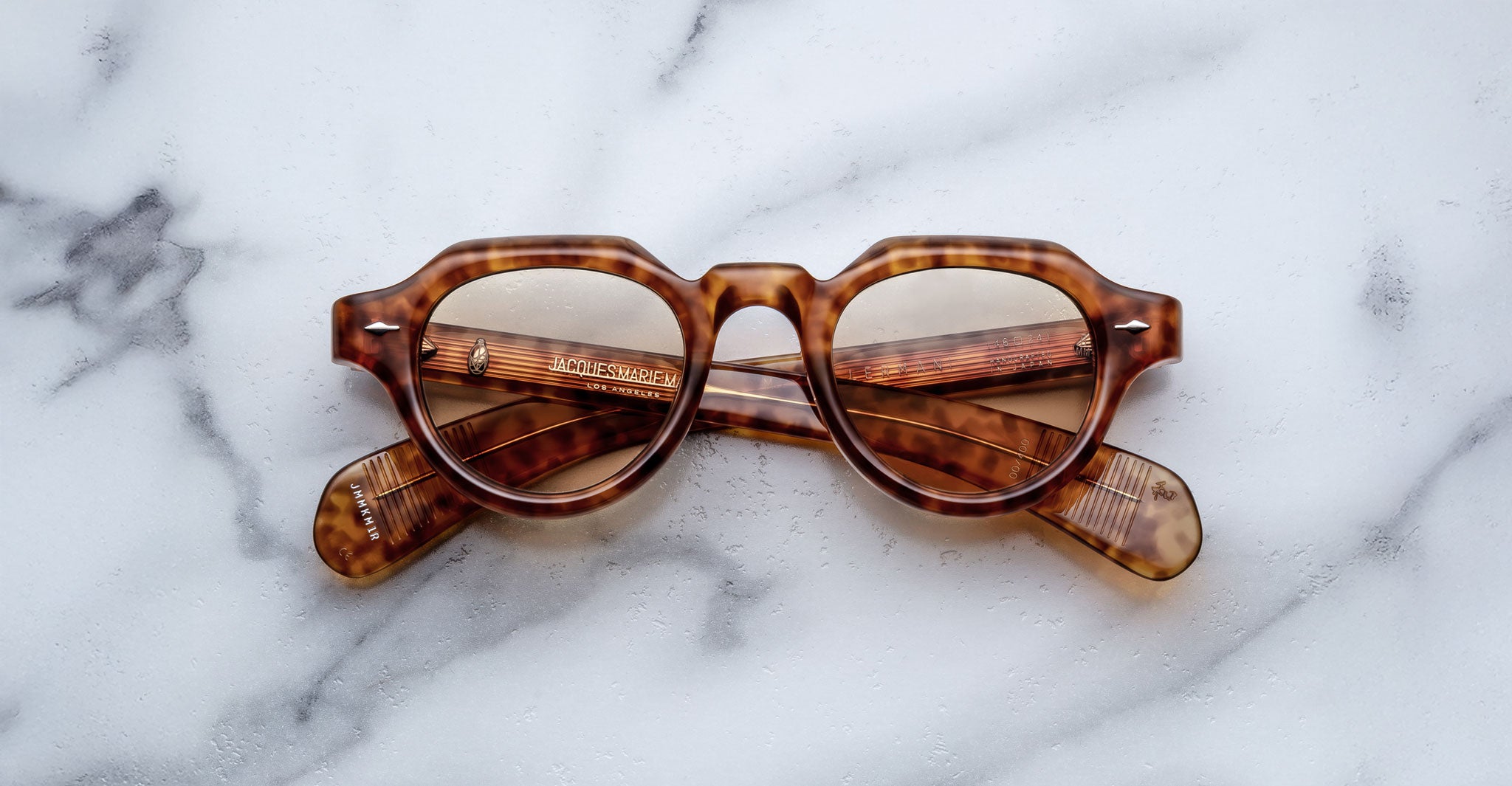 Jacques Marie Mage Kellerman brown tortoiseshell sunglasses with round lenses are folded and resting on a white marble surface, with the arms visible through the lenses.