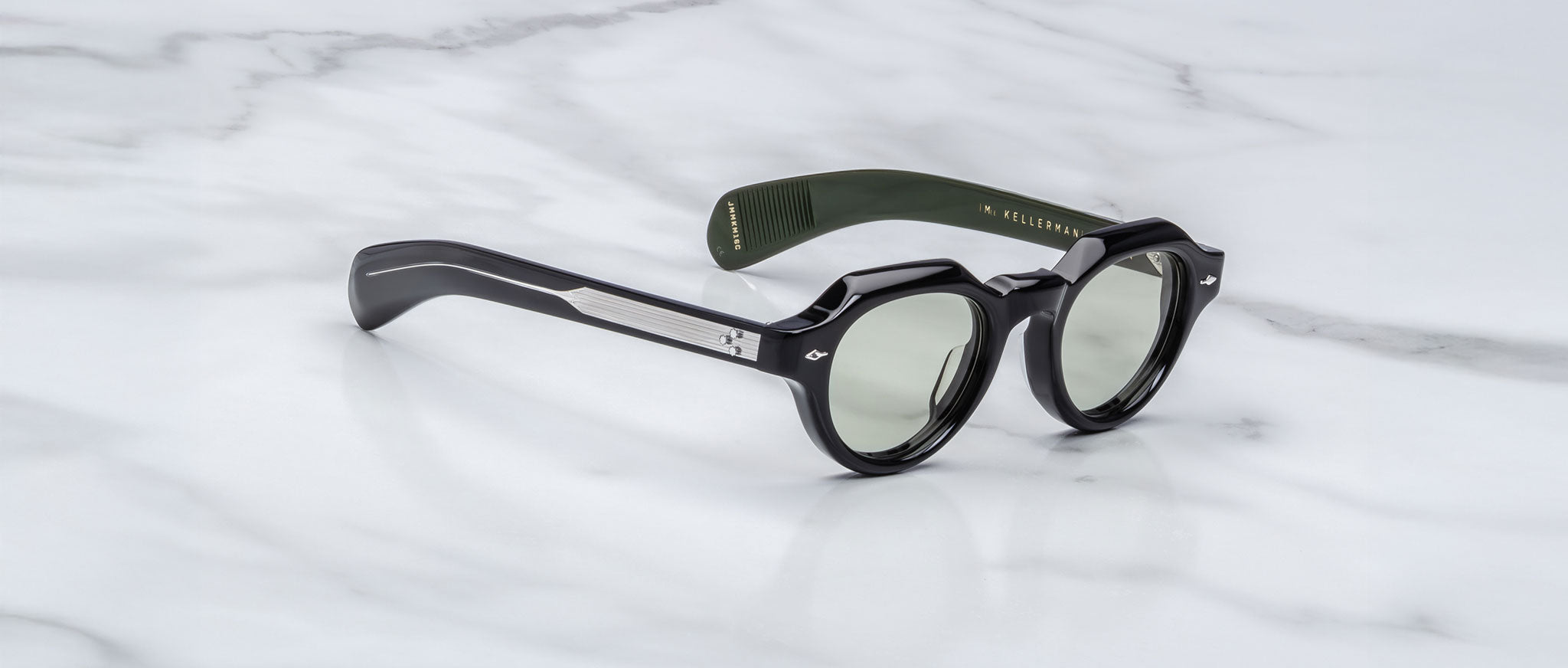 A pair of Jacques Marie Mage Kellerman sunglasses with black round lenses, silver arm details, and a green inner arm featuring gold text, resting on a white marble surface.