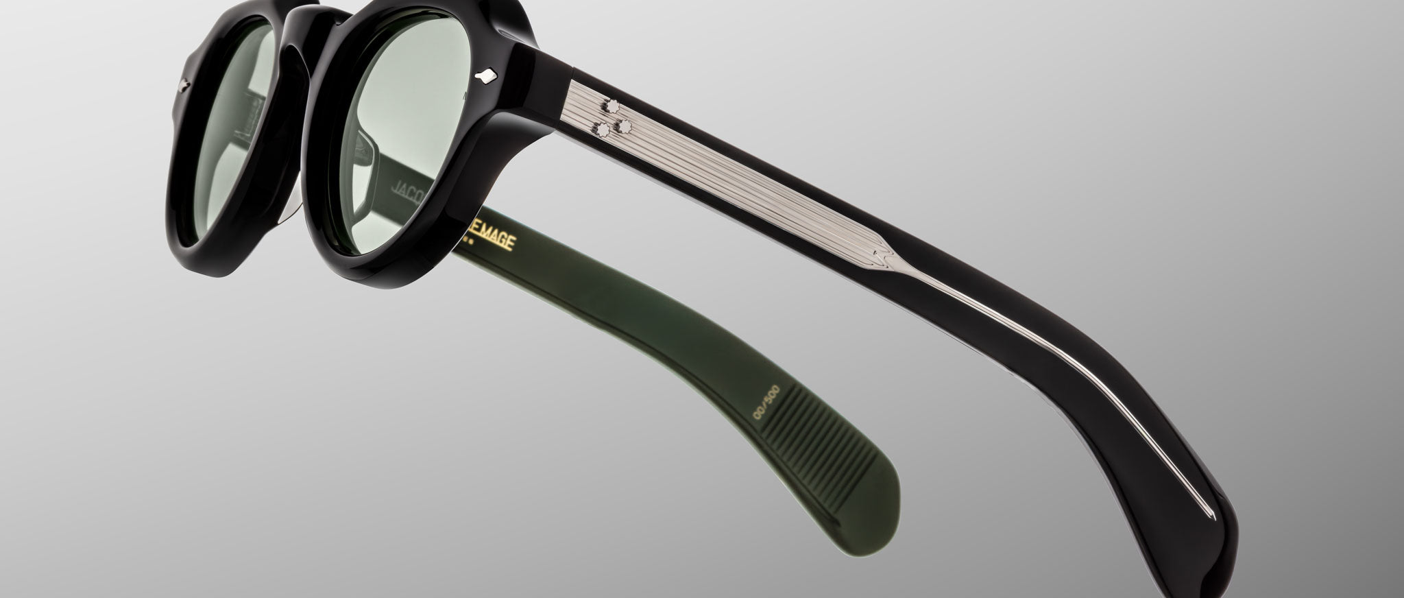 A close-up of Jacques Marie Mage Kellerman black round sunglasses with green lenses, metallic temple detailing, and an olive green inner arm with gold branding, set against a plain gray background.