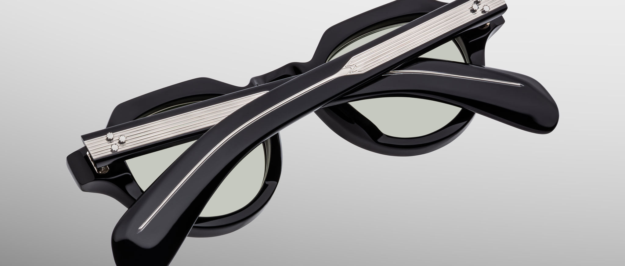 The Jacques Marie Mage Kellerman sunglasses, black and round with green-tinted lenses, are shown folded closed with crossed arms in front of the frames on a light gray background.