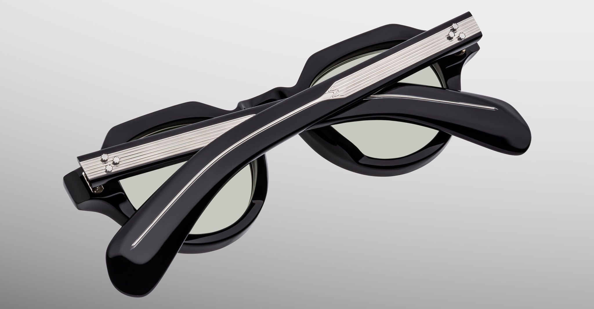 The Jacques Marie Mage Kellerman sunglasses, black and round with green-tinted lenses, are shown folded closed with crossed arms in front of the frames on a light gray background.