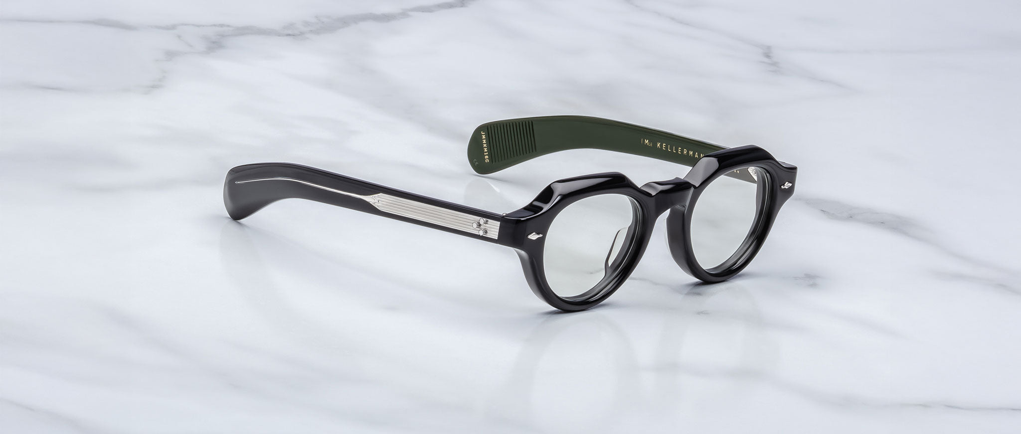 Jacques Marie Mage Kellerman black round eyeglasses with thick frames and green inner arms rest on white marble. One arm is labeled inside, while the temples display a metal accent.