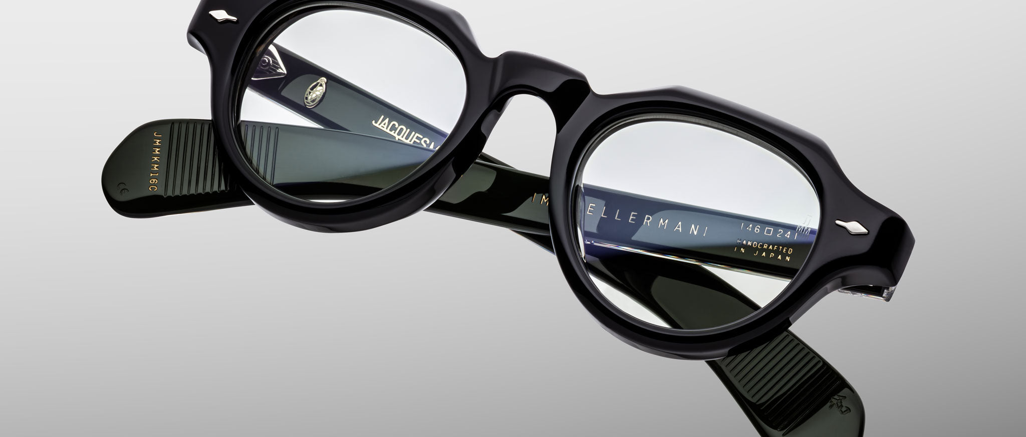 A pair of Jacques Marie Mage Kellerman black, thick-rimmed eyeglasses with round lenses is displayed on a reflective surface, revealing detailed branding and product information inside the arms.
