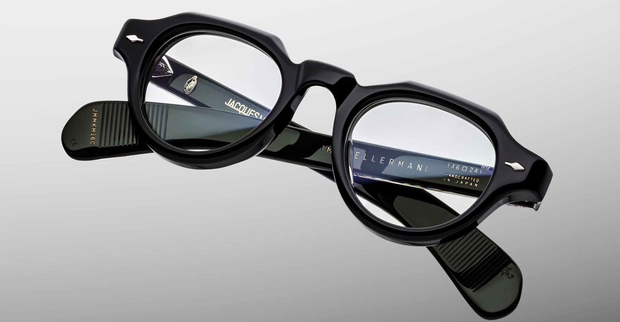 A pair of Jacques Marie Mage Kellerman black, thick-rimmed eyeglasses with round lenses is displayed on a reflective surface, revealing detailed branding and product information inside the arms.