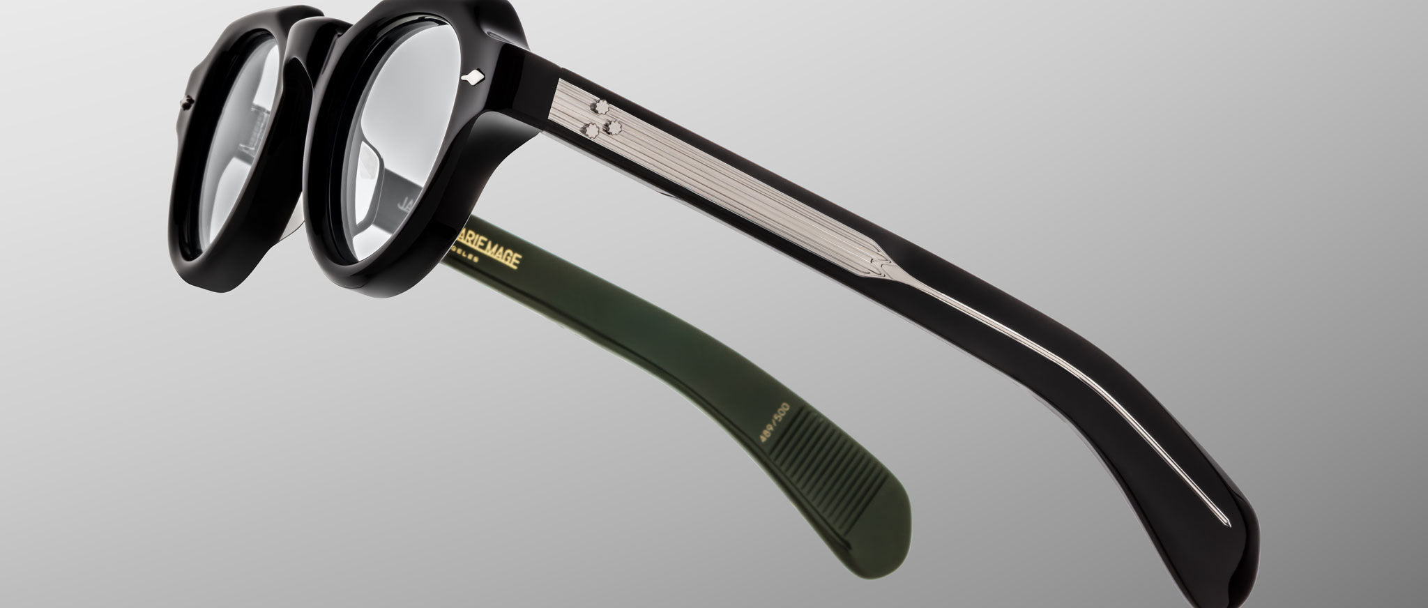 A close-up of Jacques Marie Mage Kellerman black round eyeglasses featuring silver temple detailing and one green temple tip, set against a light gray gradient background.