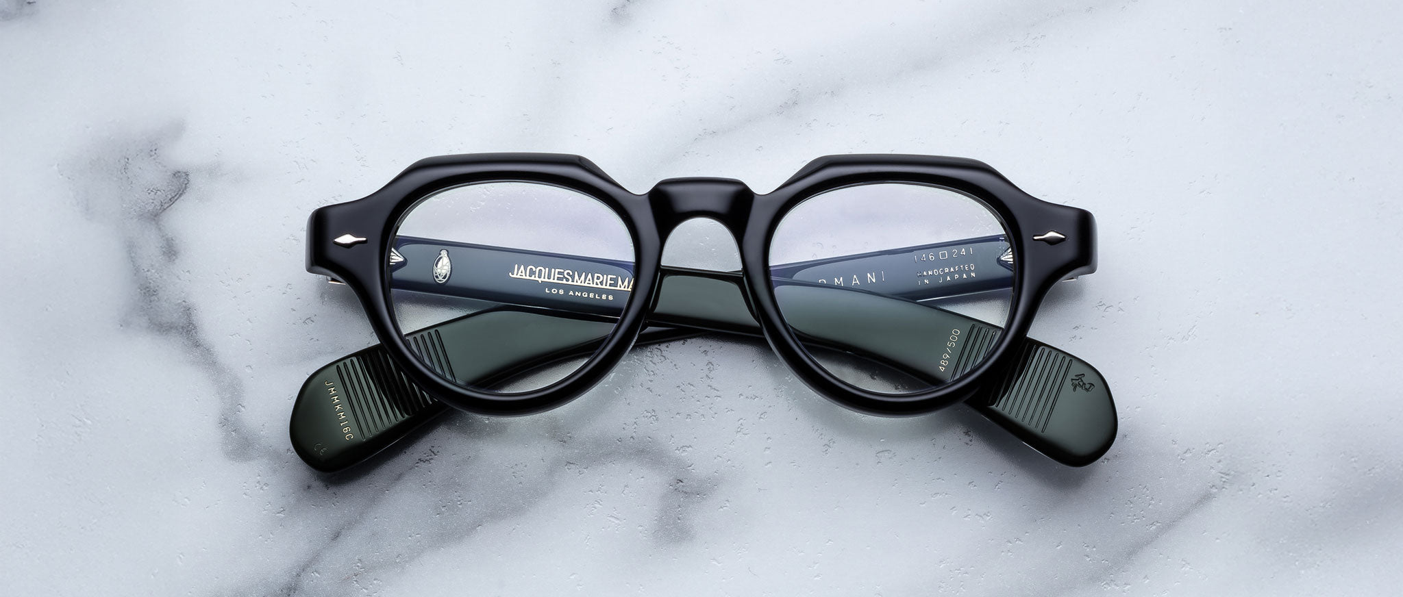 A pair of Jacques Marie Mage Kellerman black, round eyeglasses rests on a white marble surface, with brand details in white text visible on the inner arms through the clear lenses.