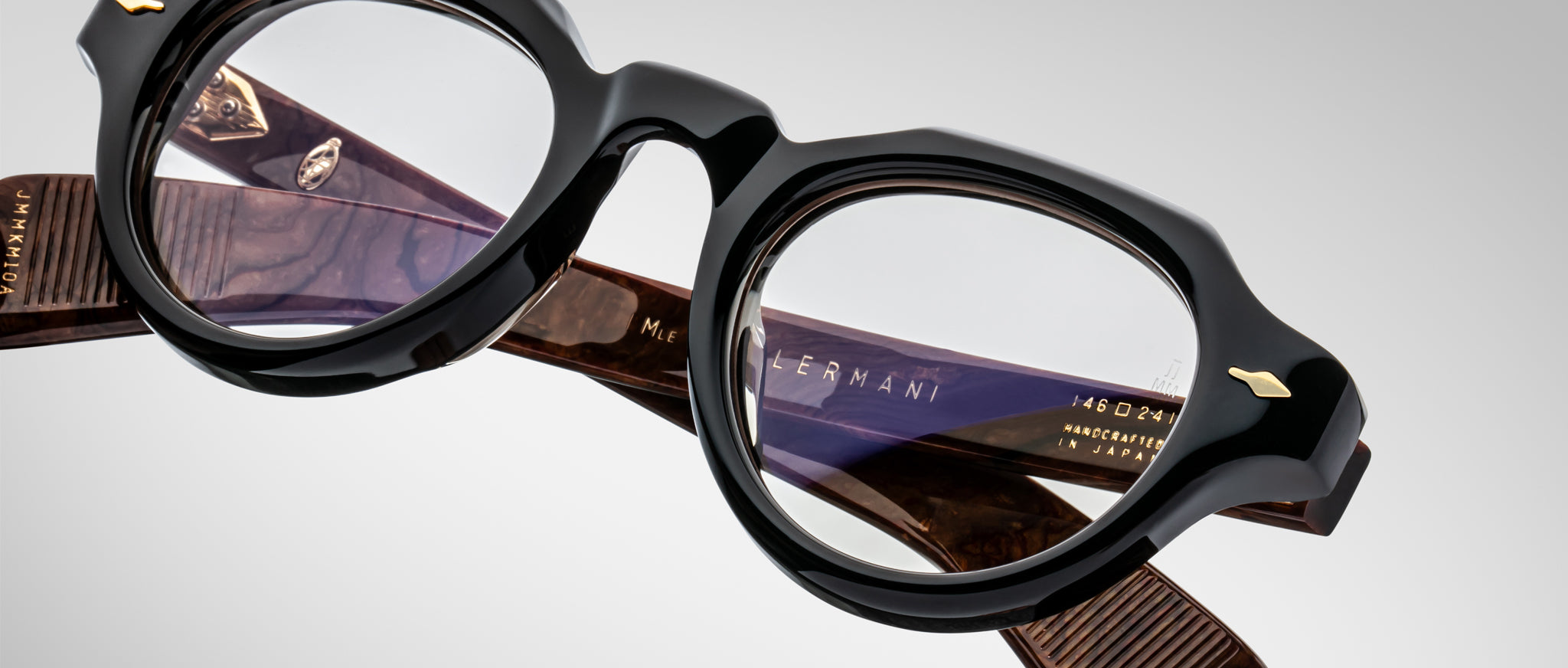 A close-up of Jacques Marie Mage Kellerman eyeglasses in black and brown shows thick frames, clear lenses, detailed hinges, gold accents, and engraved text on the arms.