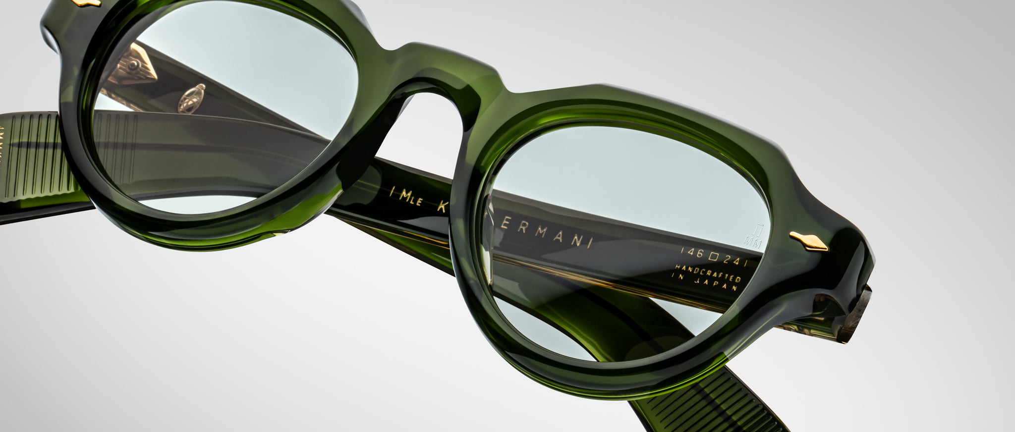Close-up of Jacques Marie Mage Kellerman chunky, translucent green eyeglasses with gold accents and engraved inner arm text, displayed on a light gray background.