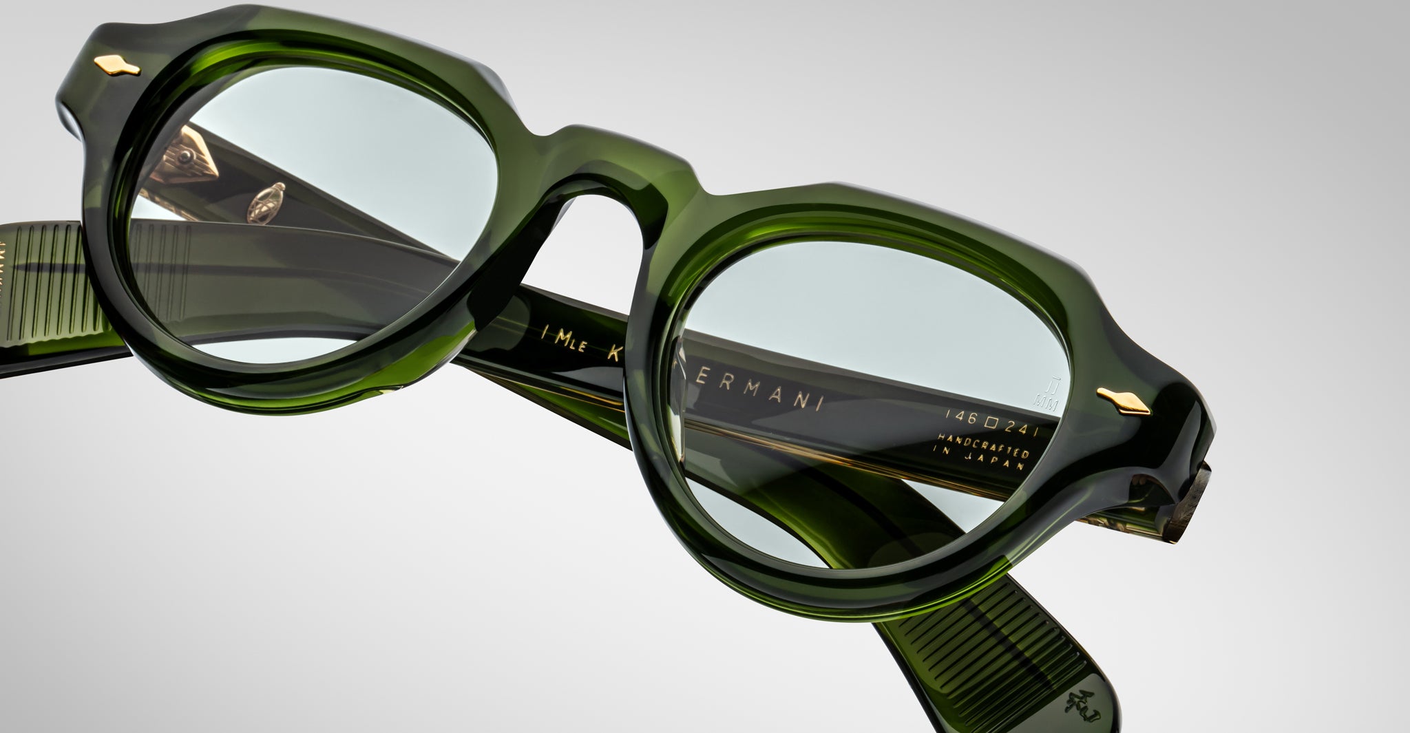 Close-up of Jacques Marie Mage Kellerman chunky, translucent green eyeglasses with gold accents and engraved inner arm text, displayed on a light gray background.