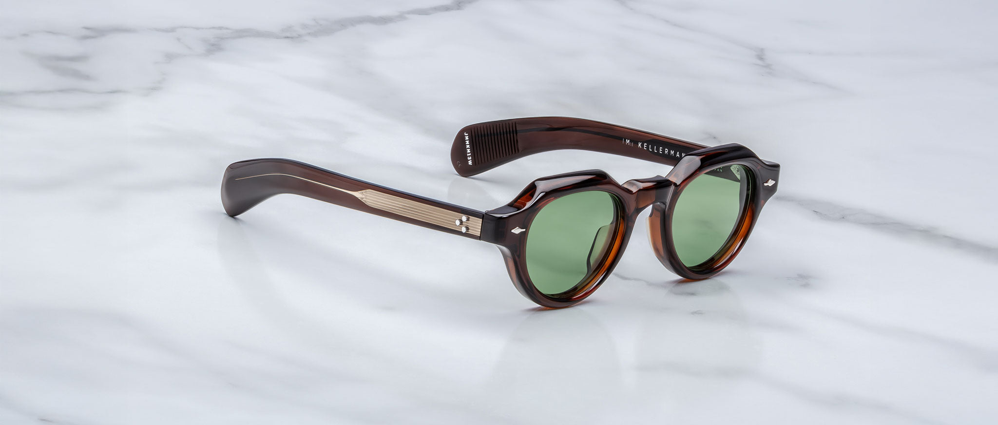 A pair of Jacques Marie Mage Kellerman sunglasses with round green lenses, thick dark brown tortoiseshell frames, and metallic arm accents rests on a white marble surface.