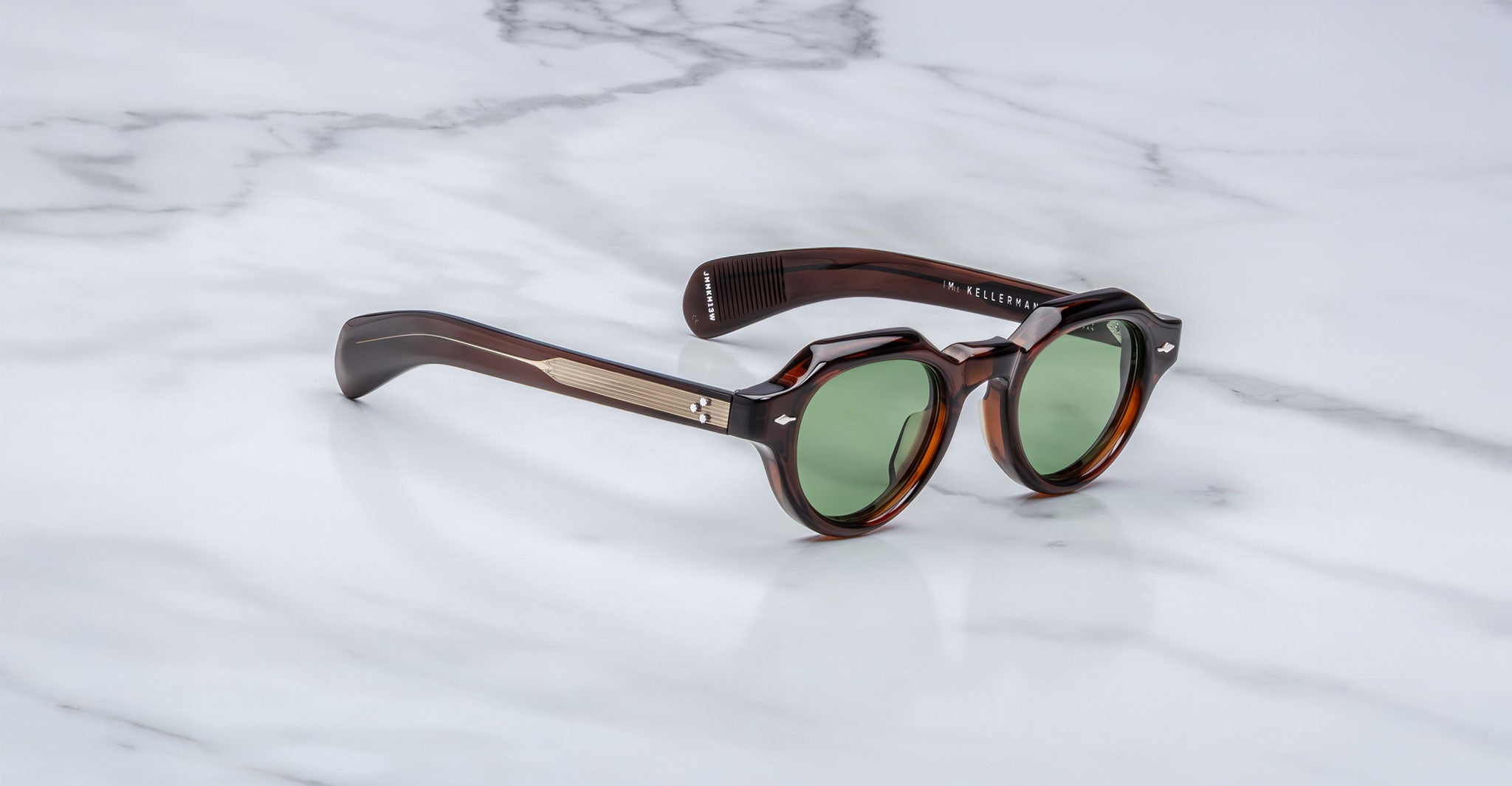A pair of Jacques Marie Mage Kellerman sunglasses with round green lenses, thick dark brown tortoiseshell frames, and metallic arm accents rests on a white marble surface.