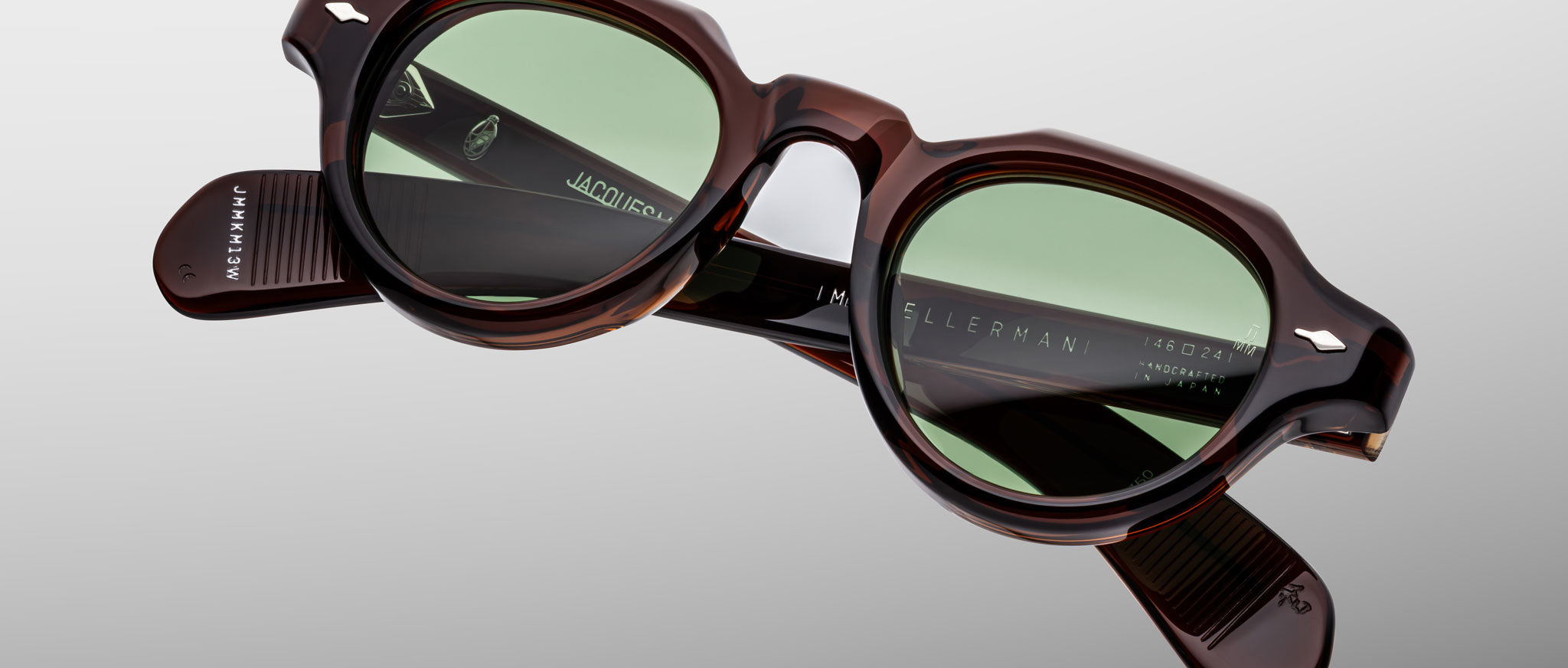 The Jacques Marie Mage Kellerman sunglasses feature round green-tinted lenses and thick tortoiseshell frames, displayed against a plain light gray background. The brand name is visible on the inside of the arms.