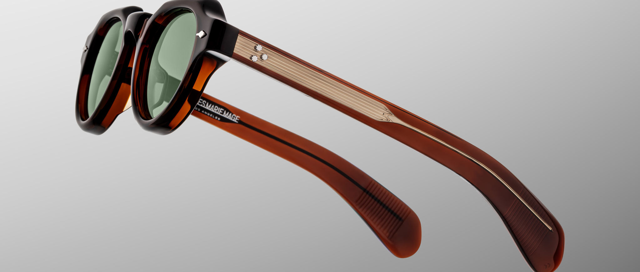 The Jacques Marie Mage Kellerman sunglasses feature dark green lenses, black and translucent brown frames, and gold-accented arms, displayed at an angle on a plain gray background.