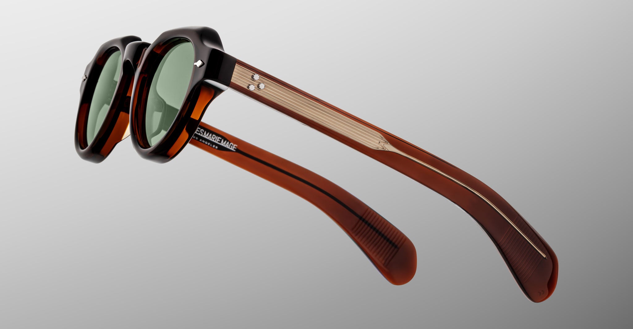 The Jacques Marie Mage Kellerman sunglasses feature dark green lenses, black and translucent brown frames, and gold-accented arms, displayed at an angle on a plain gray background.