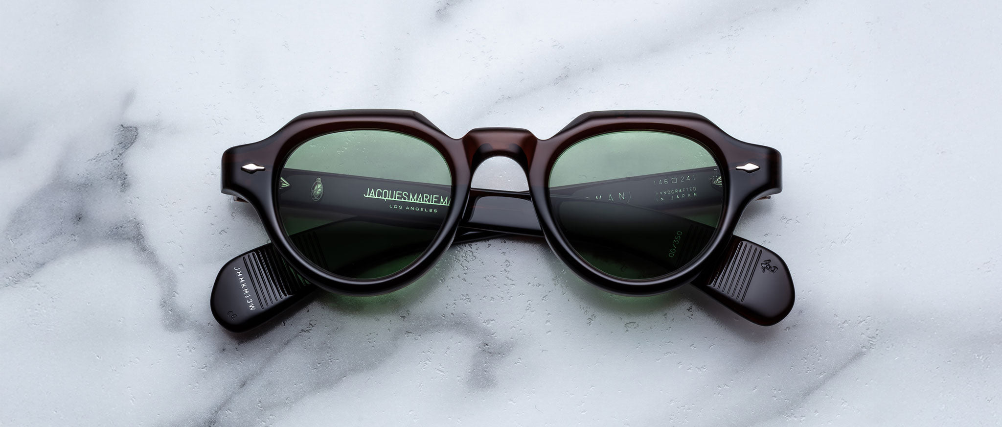 The Jacques Marie Mage Kellerman is a pair of black, thick-framed sunglasses with green tinted lenses displayed on a white marble surface.