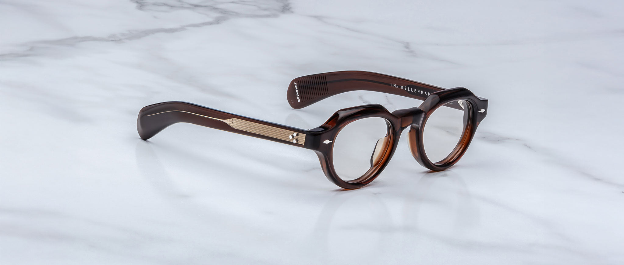 The Jacques Marie Mage Kellerman features dark brown, round eyeglasses with thick frames and metal temple accents, displayed on a white marble surface.