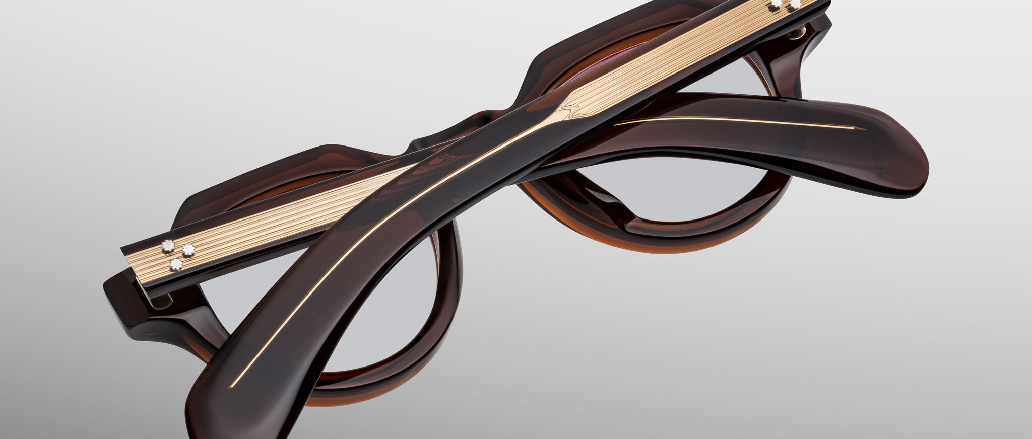 The Jacques Marie Mage Kellerman eyeglasses are shown folded, featuring thick dark brown frames with gold-toned metal temple accents against a matte gradient background.