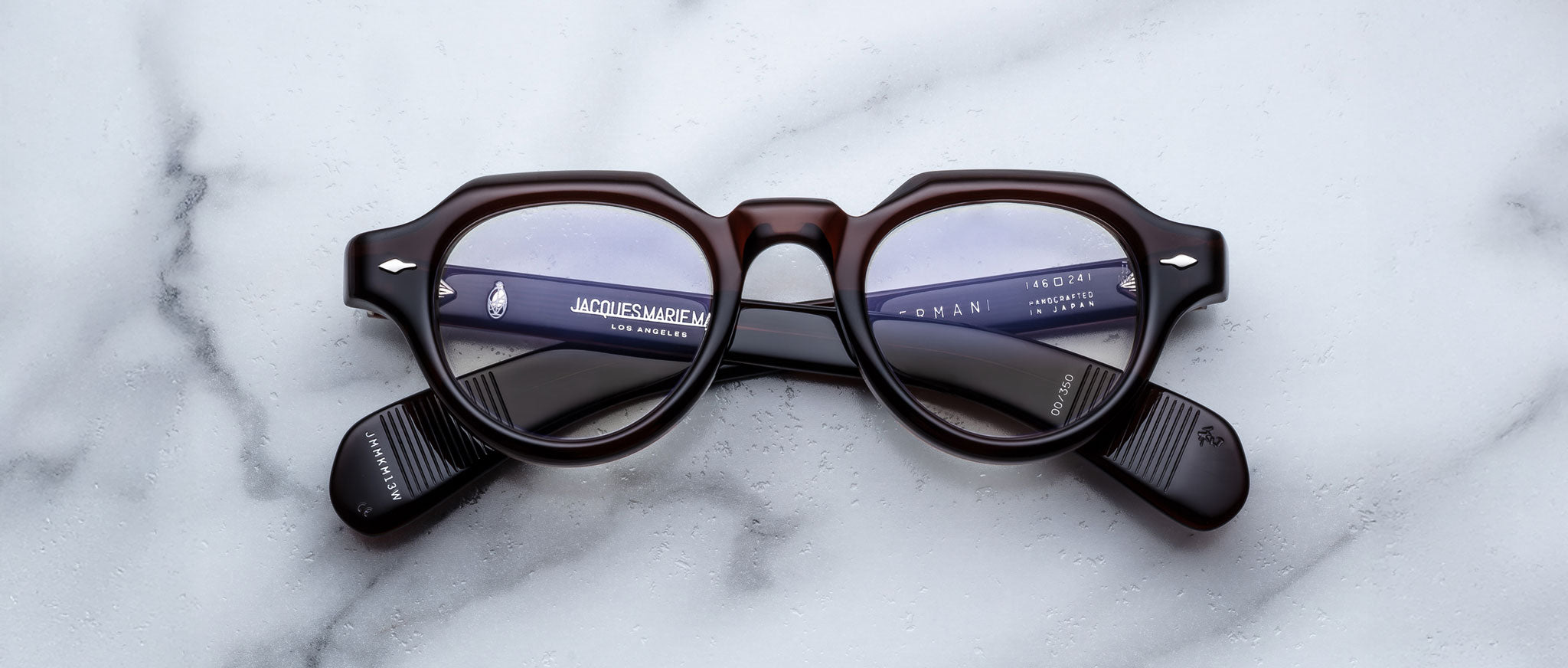 A pair of Jacques Marie Mage Kellerman round, dark-framed eyeglasses rests on a white marble surface, with text and part of a keyboard reflected in the lenses, hinting they face a computer screen.