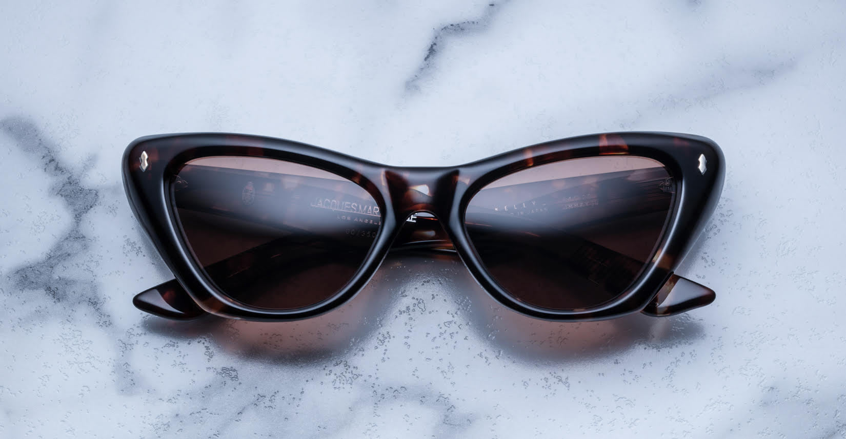 The Jacques Marie Mage Kelly cat-eye sunglasses with a dark tortoiseshell frame and tinted lenses rest on a white marble surface, with the temples partially visible.