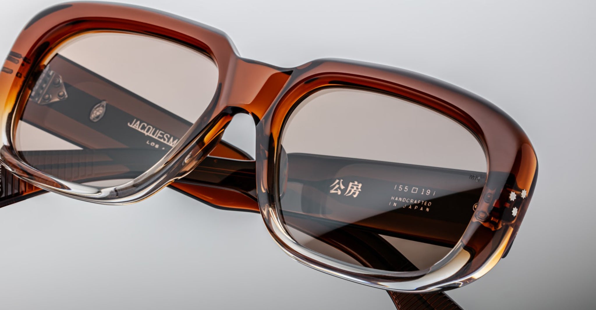 Close-up of large, rectangular Jacques Marie Mage Kobo eyeglasses in brown tint with transparent edges. The inside arms display brand markings and symbols in white, all in a sleek, polished design.