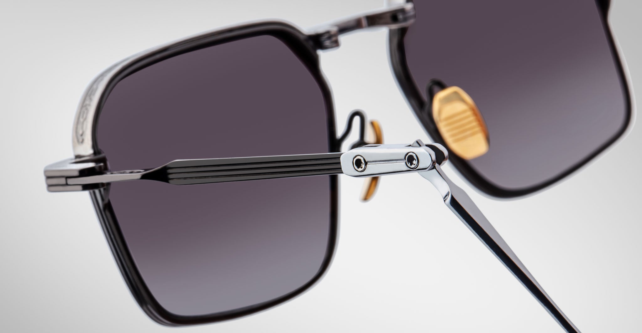 Close-up of Jacques Marie Mage Koenig sunglasses—square, black frames with dark tinted lenses, metal hinges, and gold-tone nose pads—set against a light background.