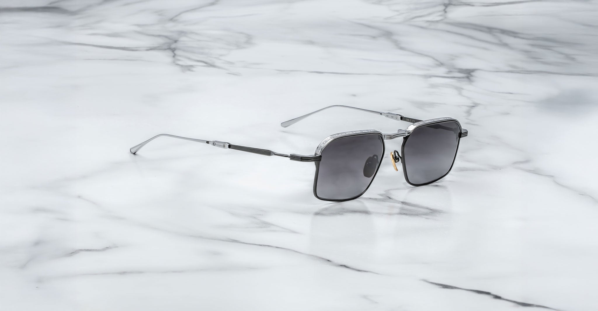 The Jacques Marie Mage Koenig features square black frames and thin metal arms, shown resting on a white marble surface with gray veining.