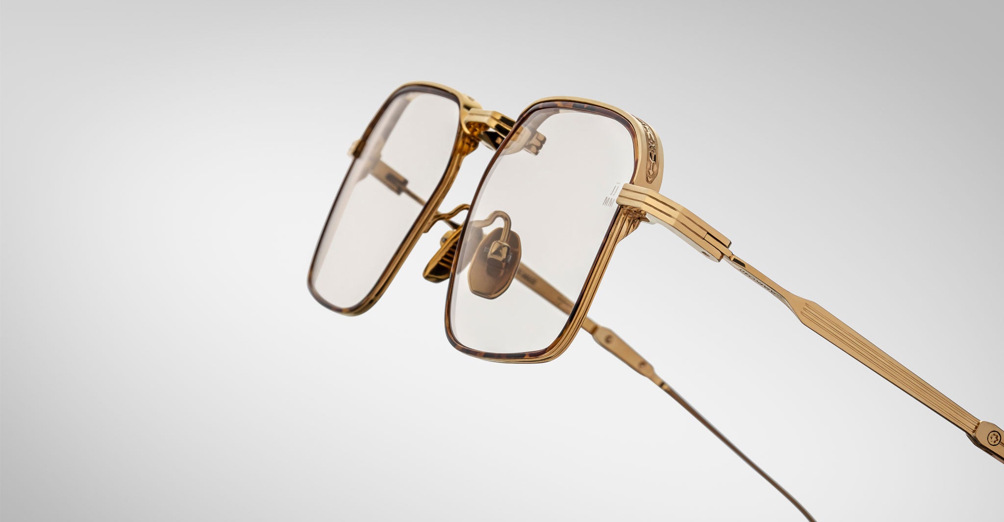 The Jacques Marie Mage Koenig eyeglasses feature gold square rims, clear lenses, textured temple detailing, and a prominent nose bridge, displayed against a white and gray gradient background.