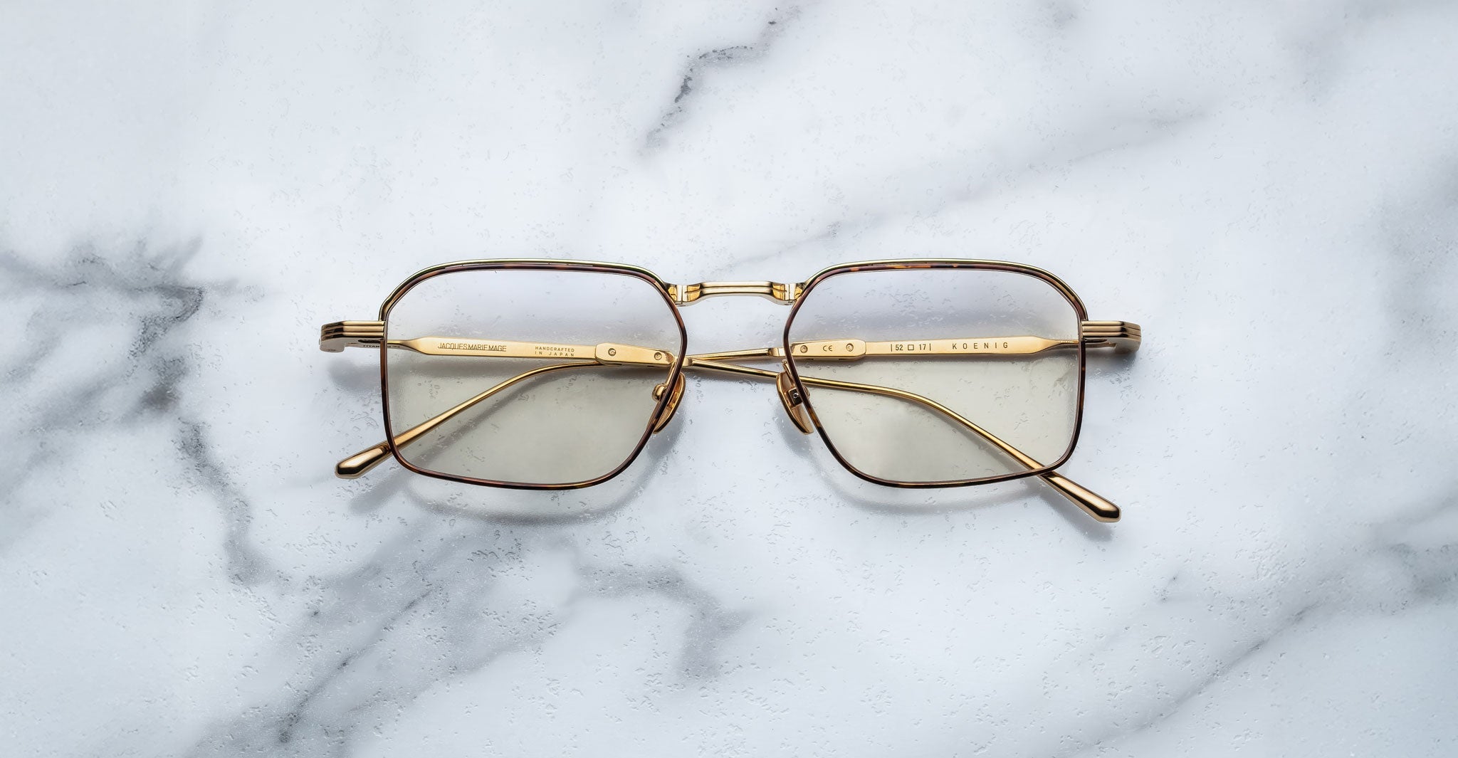 The Jacques Marie Mage Koenig eyeglasses, featuring gold rims and thin rectangular frames, are displayed with open arms facing upward on a white marble surface.