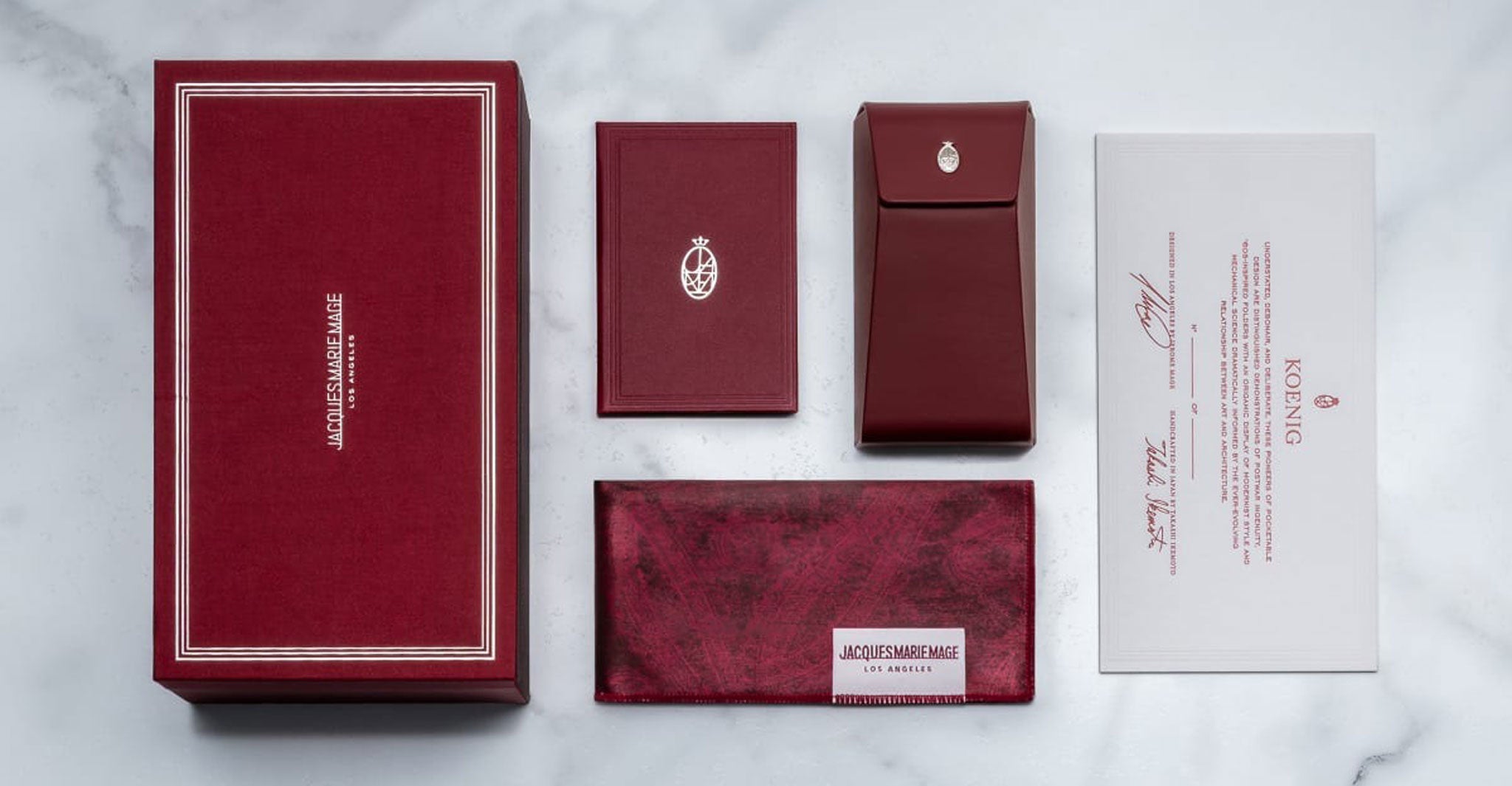 The Jacques Marie Mage Koenig set features red and white luxury packaging, including a large box, fabric pouch, card holder, envelope, and certificate—all elegantly arranged on a white marble surface.
