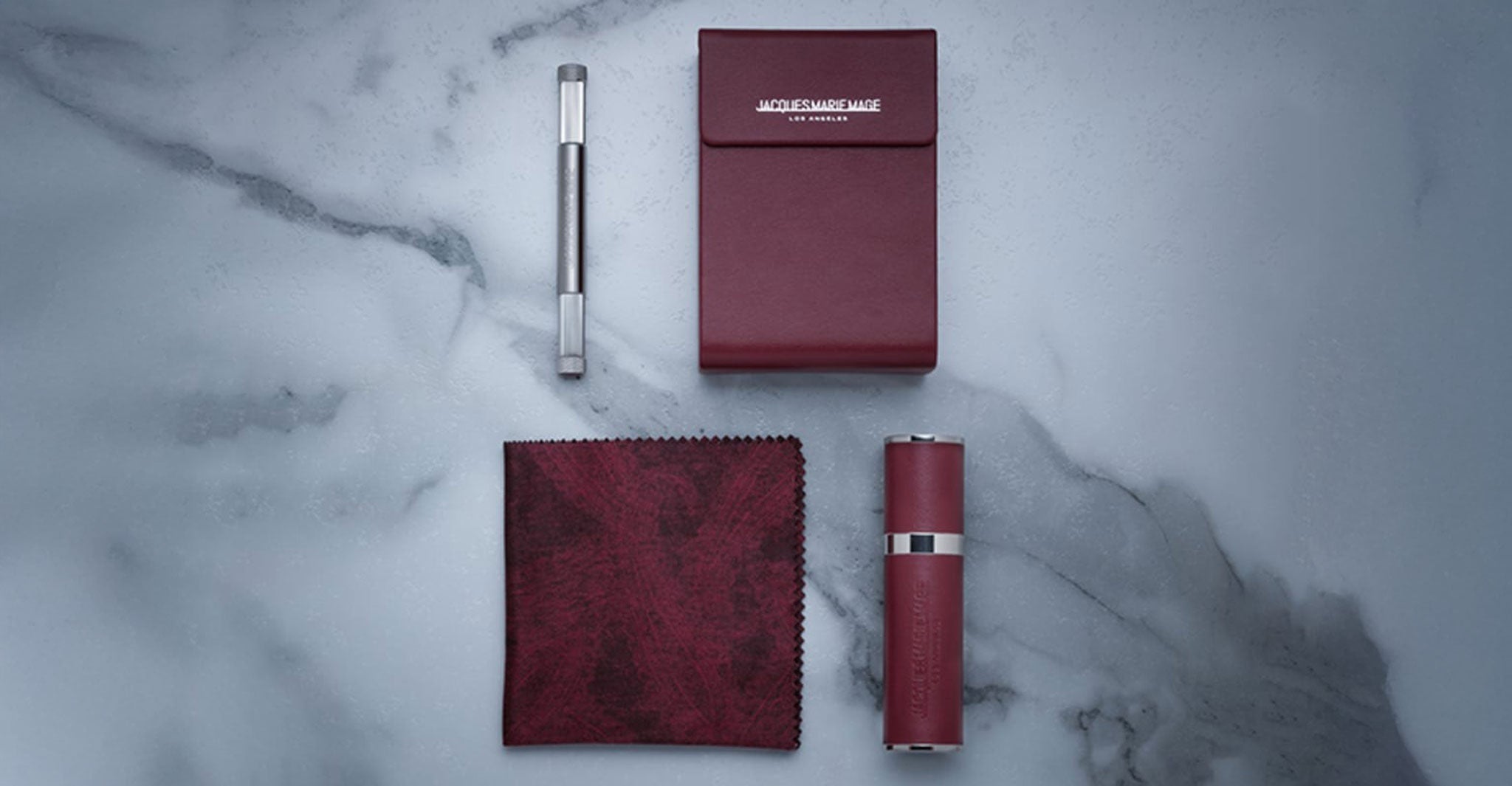 The Jacques Marie Mage Koenig set features a silver pen, a burgundy notepad, a matching cylindrical container, and a dark red cleaning cloth, all arranged neatly on a light marble surface.