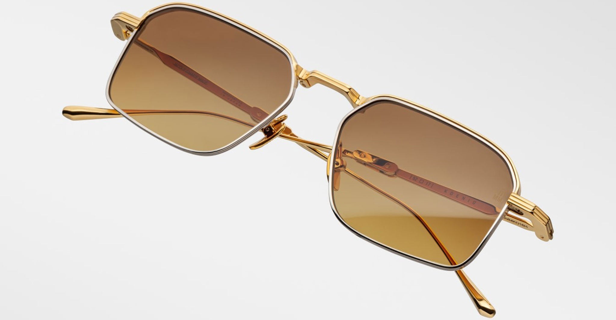 The Jacques Marie Mage Koenig sunglasses are displayed against a white background, featuring gold rectangular frames, brown tinted lenses, thin arms, and a subtle brown accent.