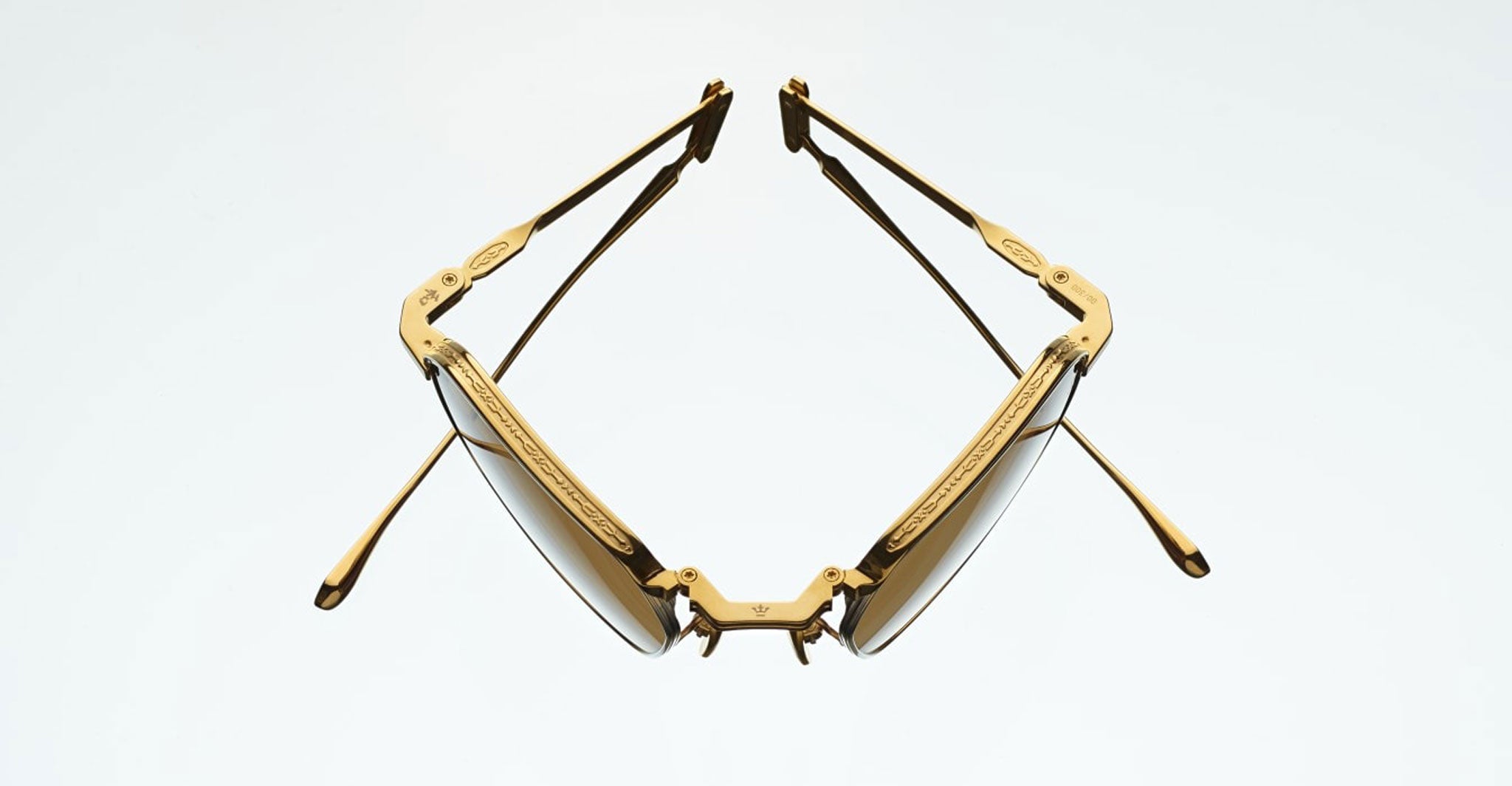 A pair of Jacques Marie Mage Koenig gold sunglasses is displayed from above with open temples, angled upward on a plain light background. The frame is accented with decorative engravings on the sides.