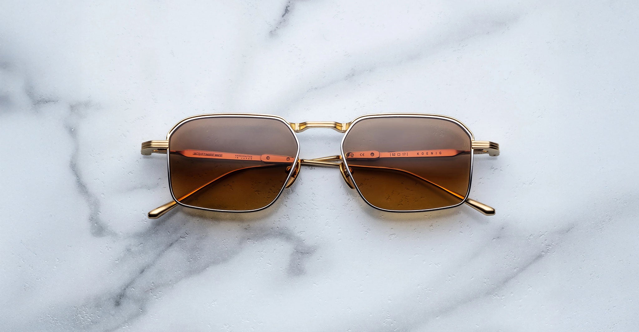 The Jacques Marie Mage Koenig sunglasses, featuring square gold frames, brown lenses, and red-and-gold arm details, rest on a white marble surface.