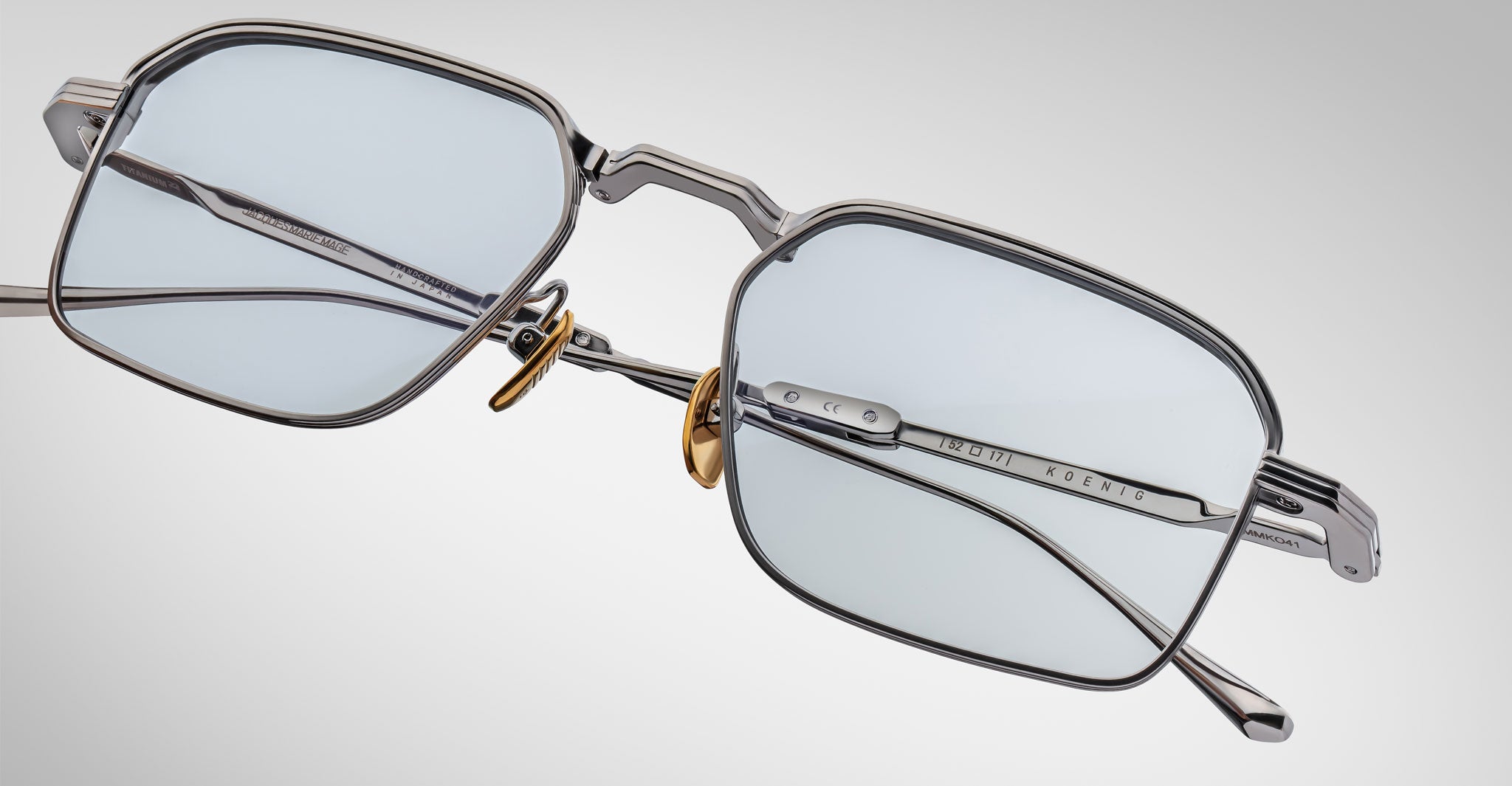 A close-up of Jacques Marie Mage Koenig silver rectangular eyeglasses with light blue-tinted lenses, gold nose pads, and visible inner temple branding set against a plain light gray background.