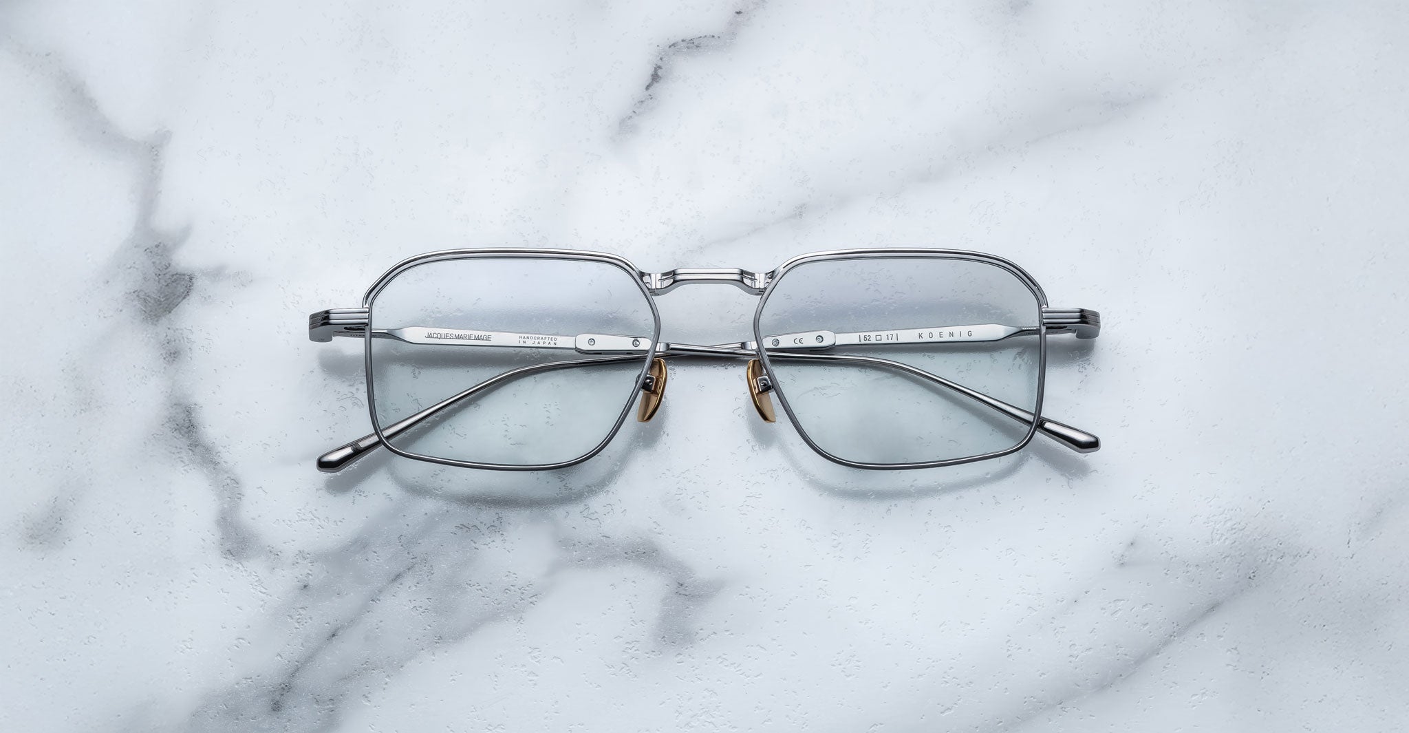 A pair of Jacques Marie Mage Koenig rectangular silver metal eyeglasses with clear lenses is displayed open on a white marble surface, temples facing upward.