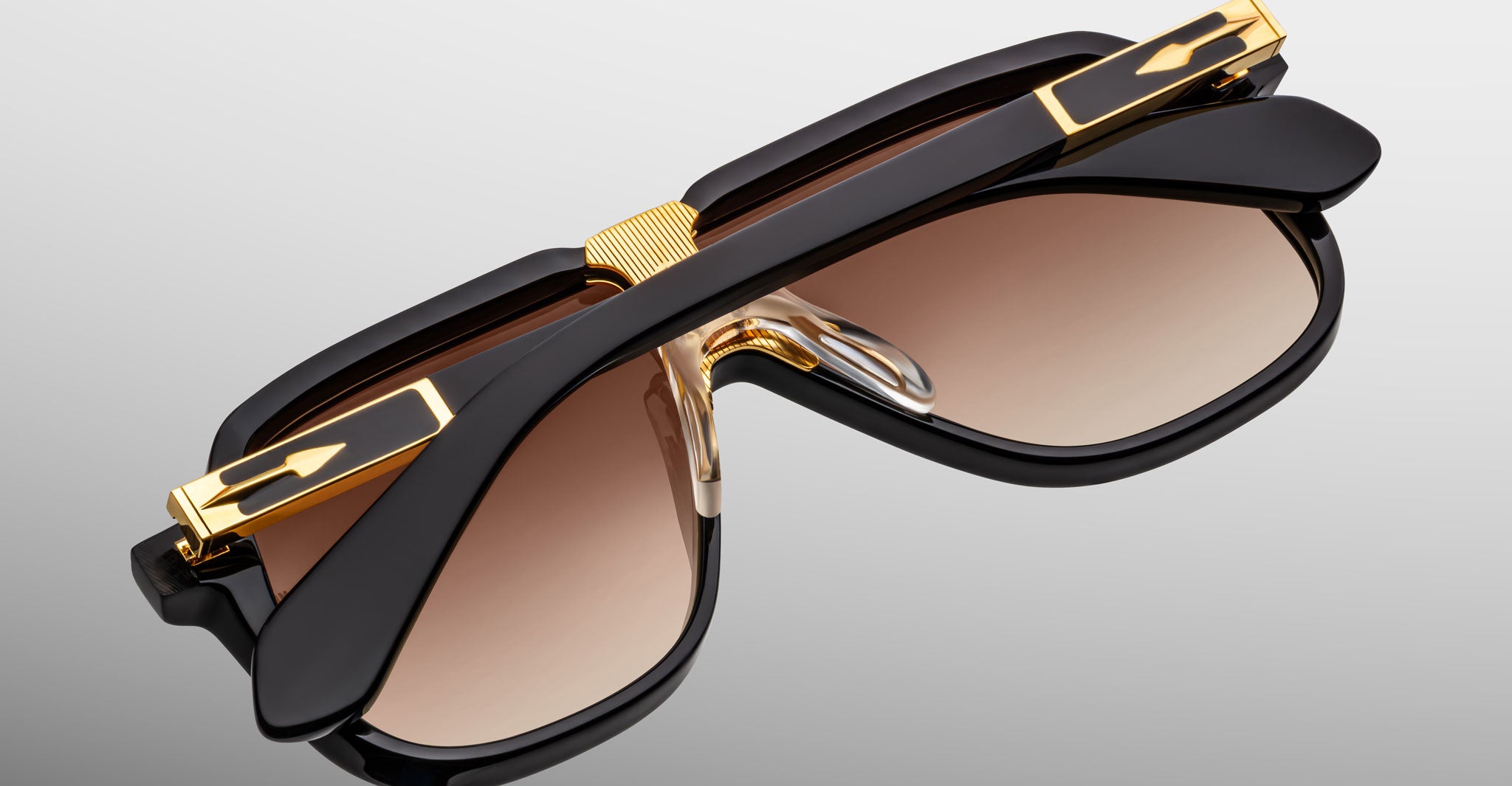 The Jacques Marie Mage Kurokawa sunglasses feature a stylish black frame with gold accents and brown gradient lenses, elegantly displayed folded on a reflective gray surface.