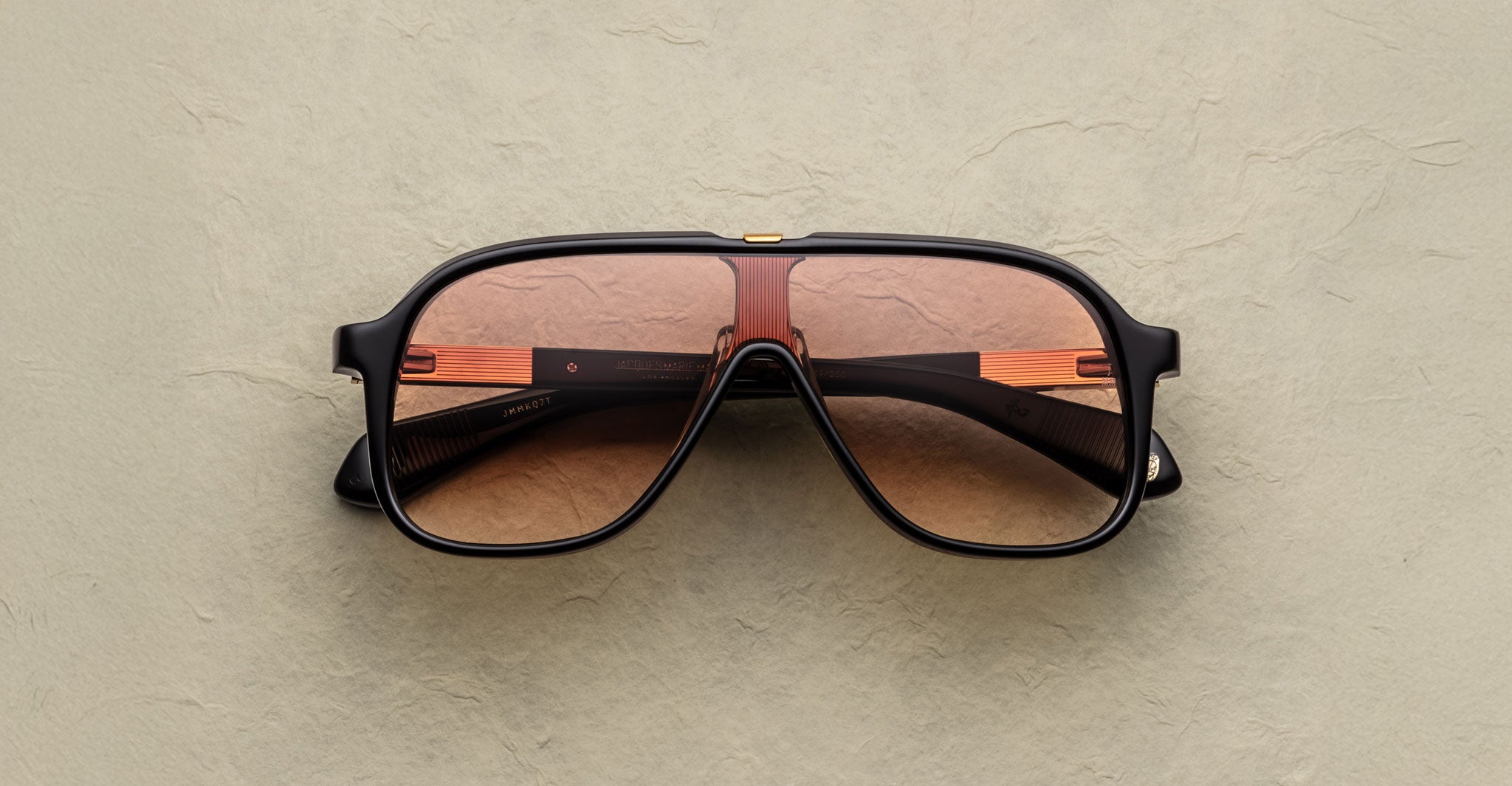 The Jacques Marie Mage Kurokawa sunglasses, featuring black frames, thick arms, and large brown-tinted lenses in a retro aviator style, rest on a textured beige surface.