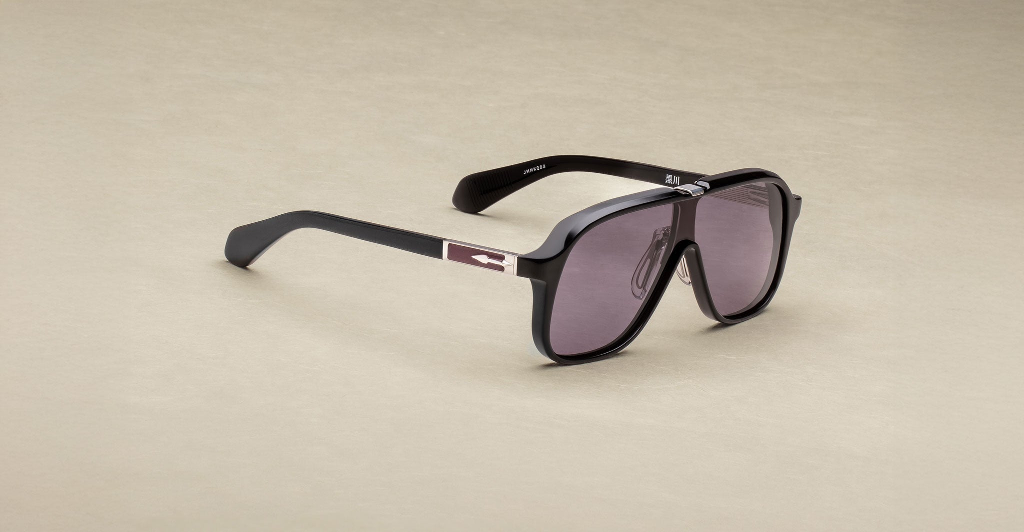 The Jacques Marie Mage Kurokawa is a pair of oversized black sunglasses with dark tinted lenses and silver arrow detailing on the temples, shown on a light beige surface.
