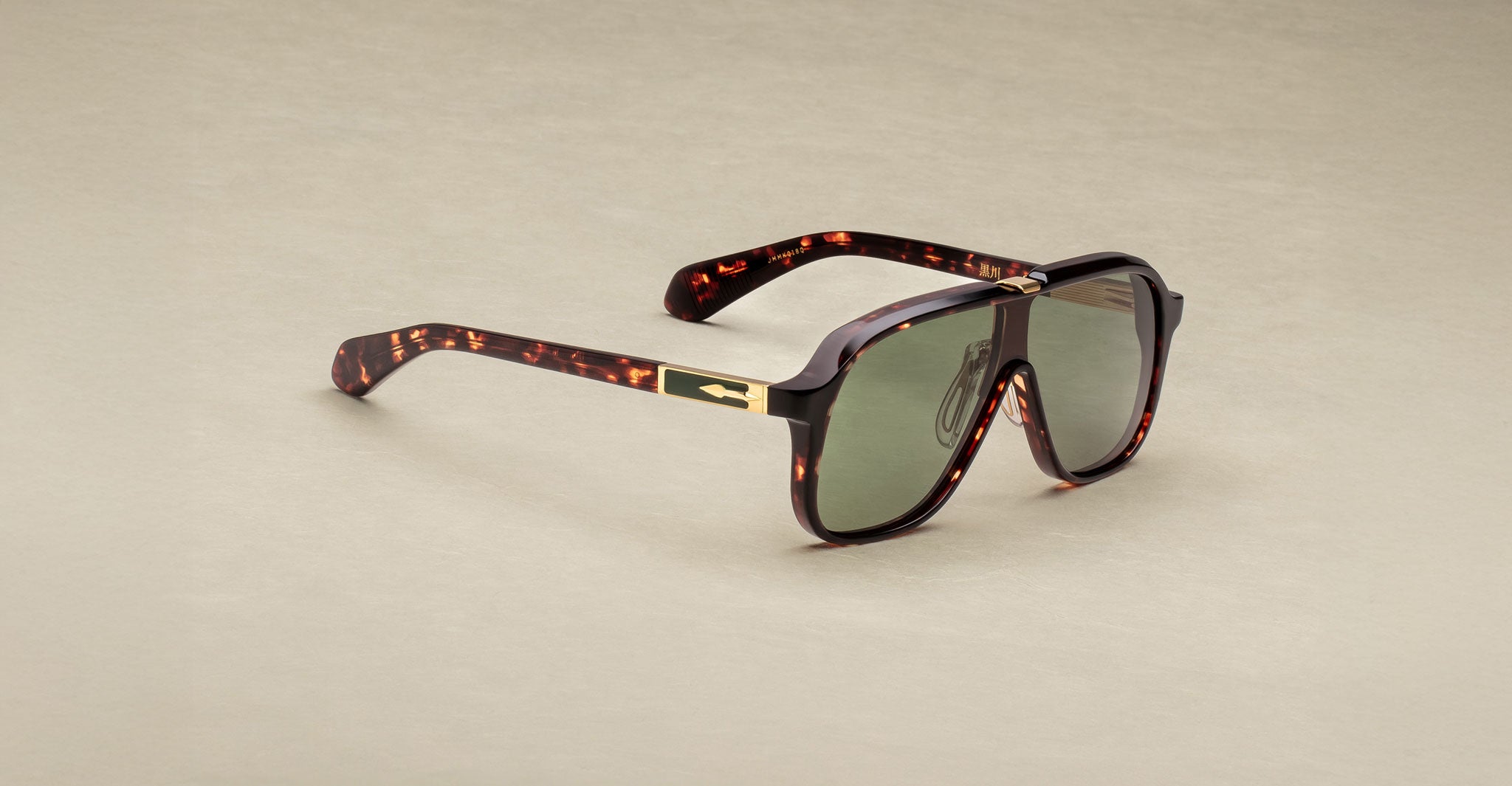 The Jacques Marie Mage Kurokawa sunglasses feature thick tortoiseshell patterned frames, green-tinted lenses, and temples with metallic accents and a subtle logo, displayed on a neutral beige surface.