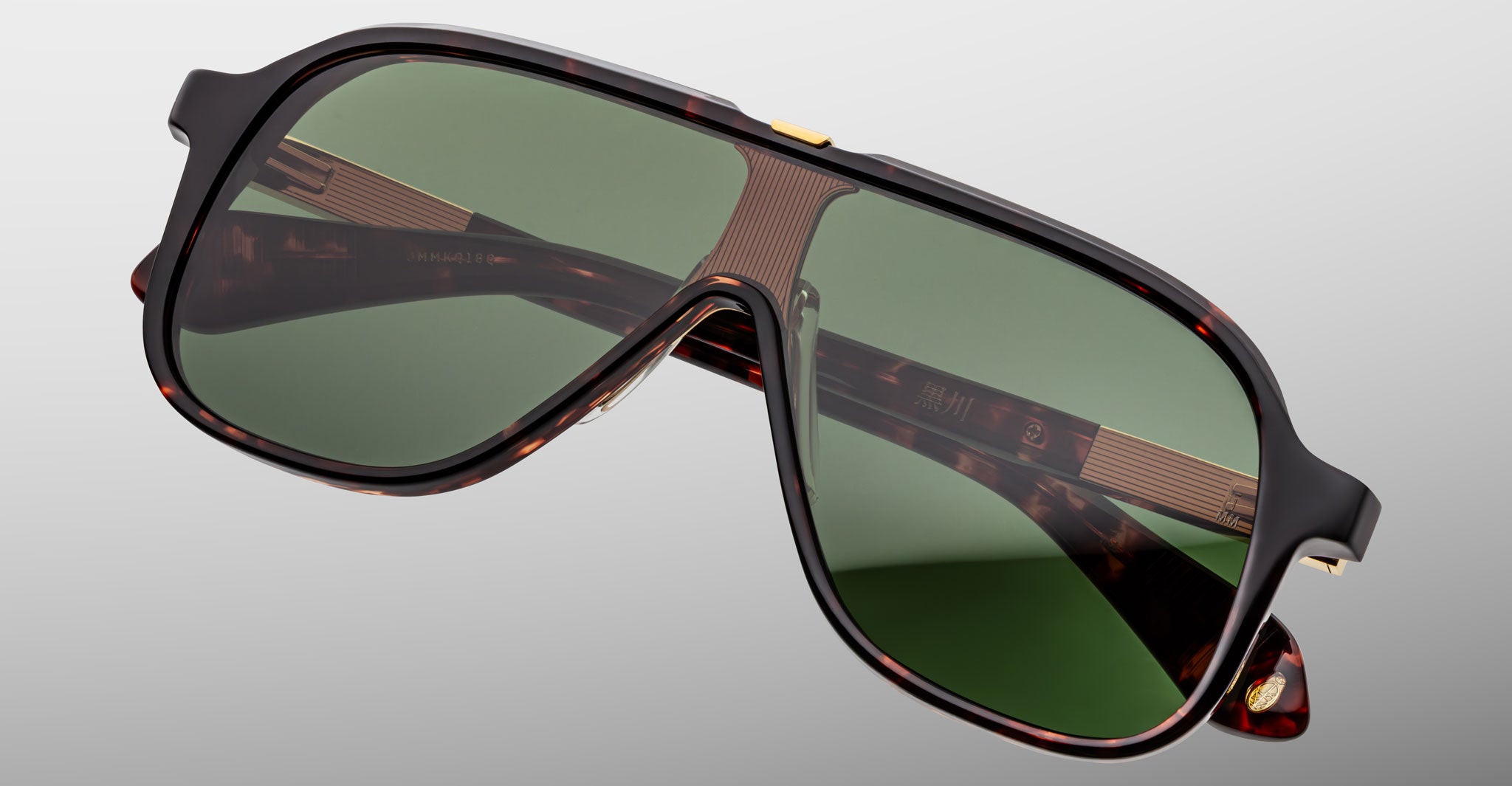 The Jacques Marie Mage Kurokawa is a pair of oversized, tortoiseshell sunglasses with green-tinted lenses and gold accents, displayed on a flat, light gray surface.
