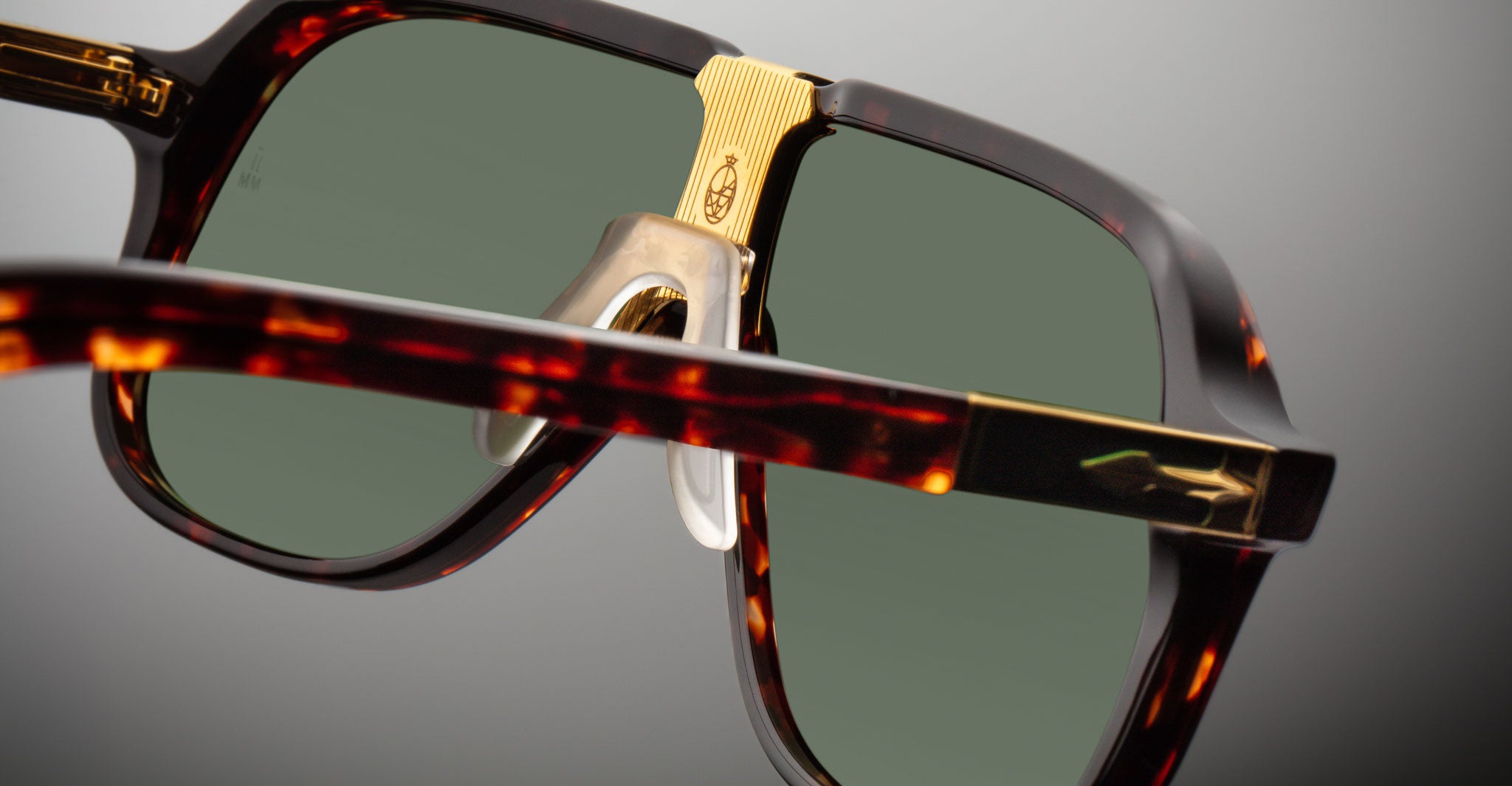 Close-up of Jacques Marie Mage Kurokawa sunglasses with tortoiseshell frames, green-tinted lenses, gold nose bridge detailing, and transparent nose pads against a plain gray background.