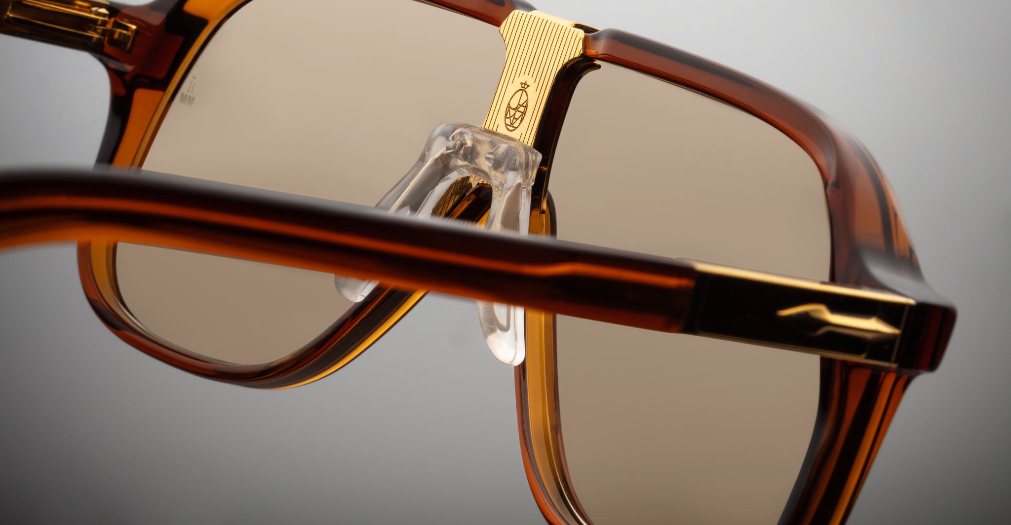 Close-up of Jacques Marie Mage Kurokawa sunglasses in brown and gold, featuring clear nose pads, a detailed gold bridge, and frame accents set against a neutral background.